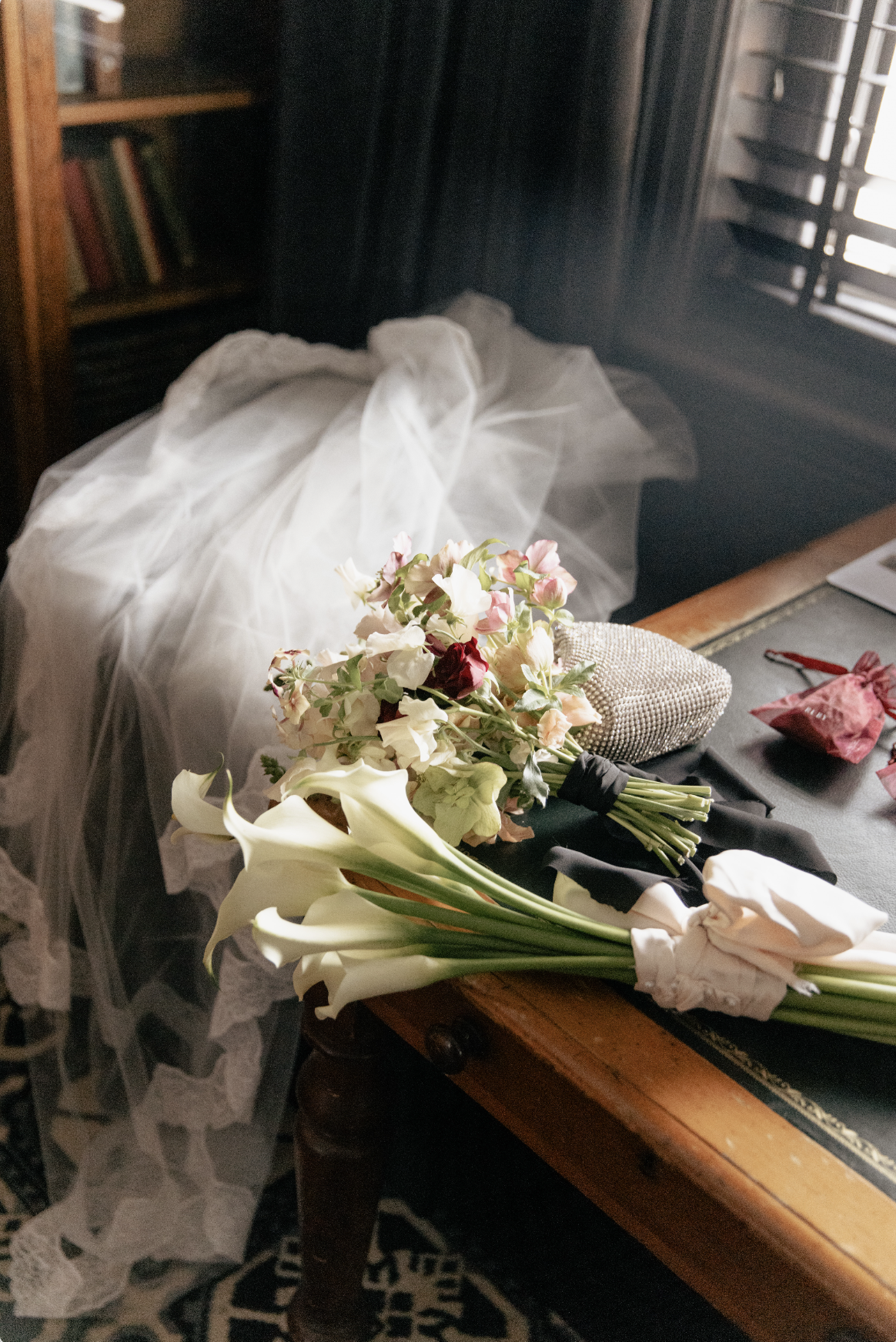 A bouquet of white, pink, and red flowers resting on a black cloth on a wooden table, with a white veil and other wedding accessories nearby, in a dimly lit room with window shutters.