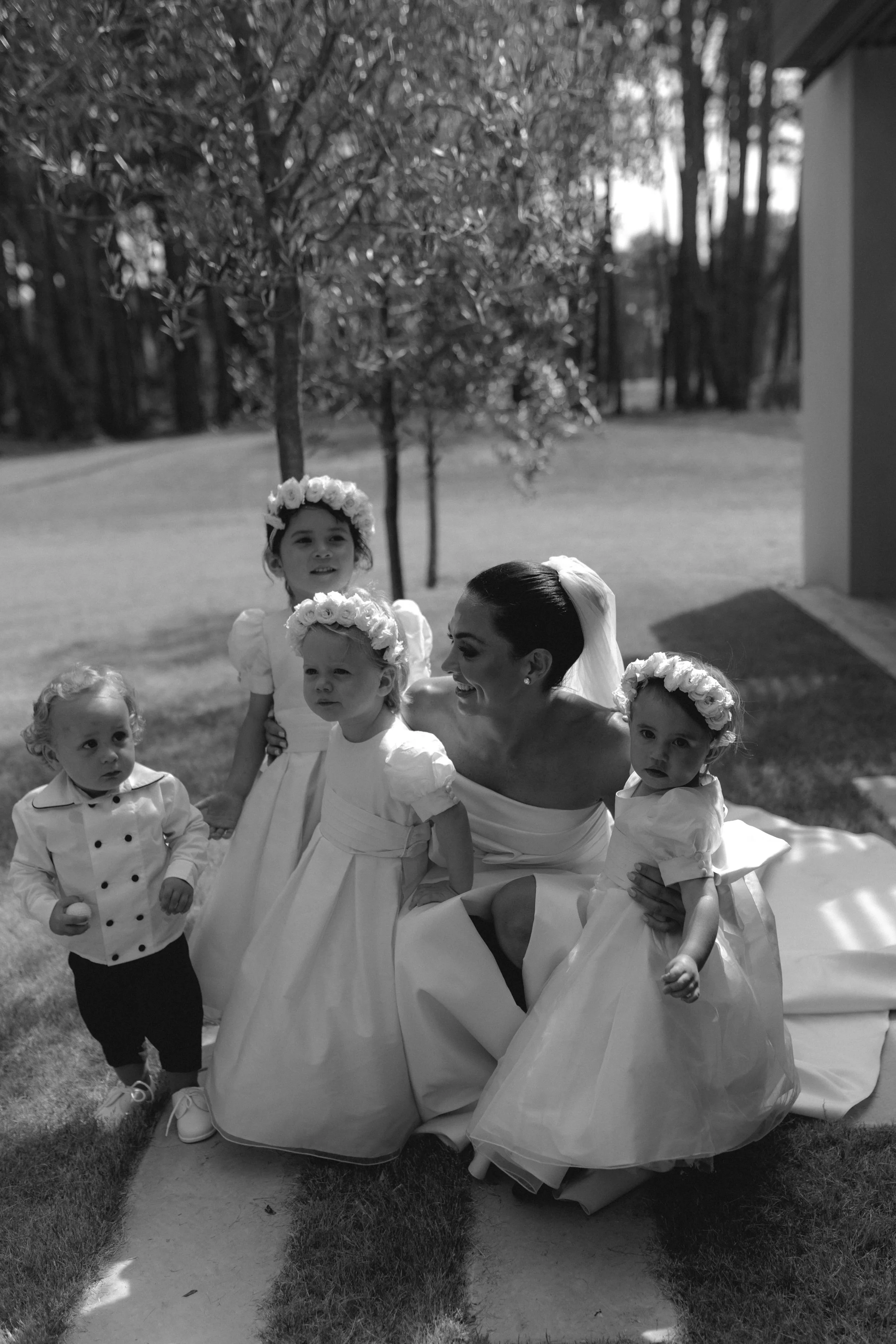 A woman in a wedding dress surrounded by five young children dressed in formal attire, outdoor setting with trees and a pathway.