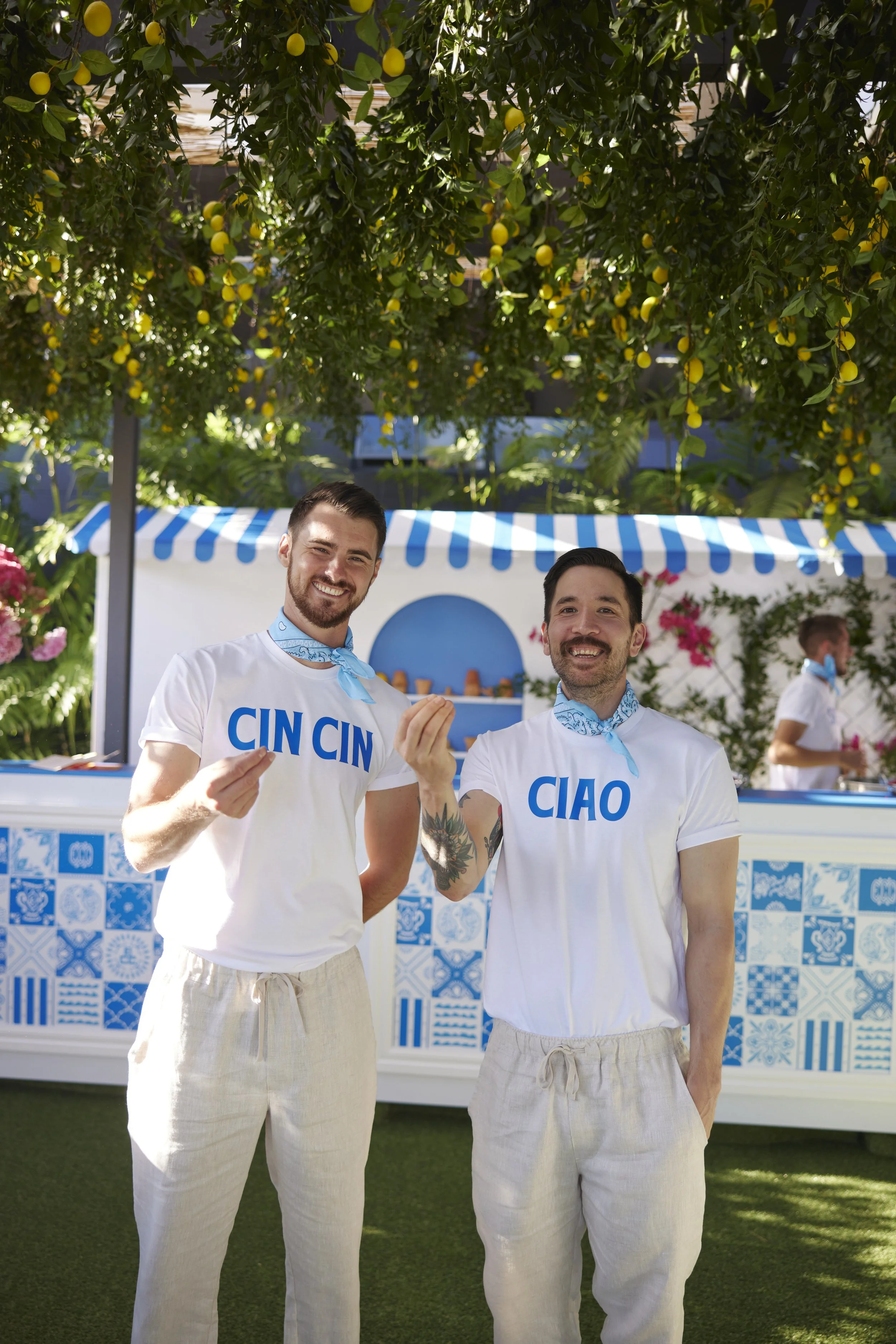Two smiling men wearing white t-shirts with blue text that says 'CIN' and 'CIAO', standing outdoors under a lemon tree. They are dressed in beige pants, with one man having a tattoo on his left arm and the other wearing a blue scarf around his neck. A stand with a blue and white striped canopy and decorative tiles are visible in the background, suggesting a festive setting.