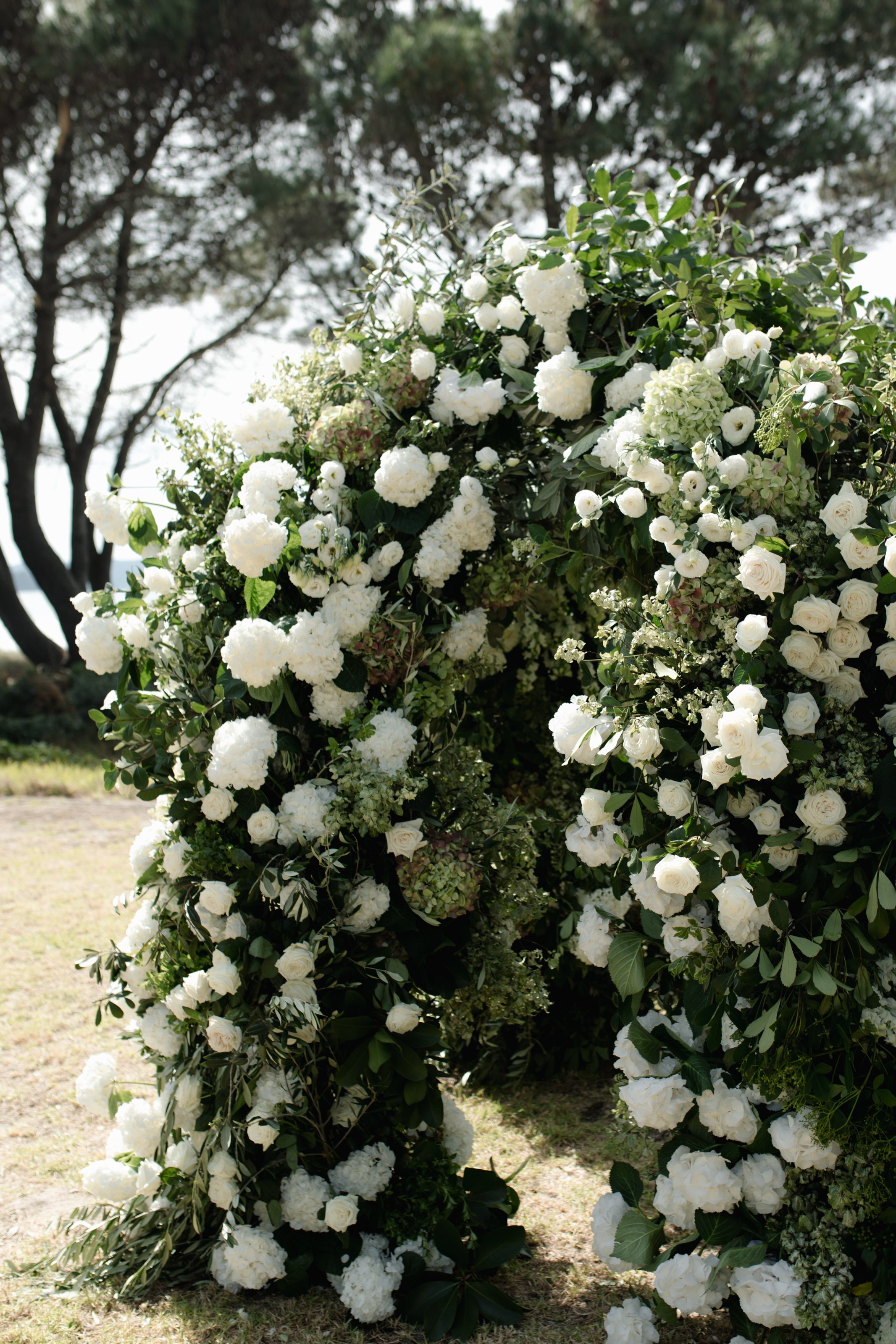 A large floral arrangement of white hydrangeas, roses, and other white flowers with green leaves, outdoors on grass with trees in the background.