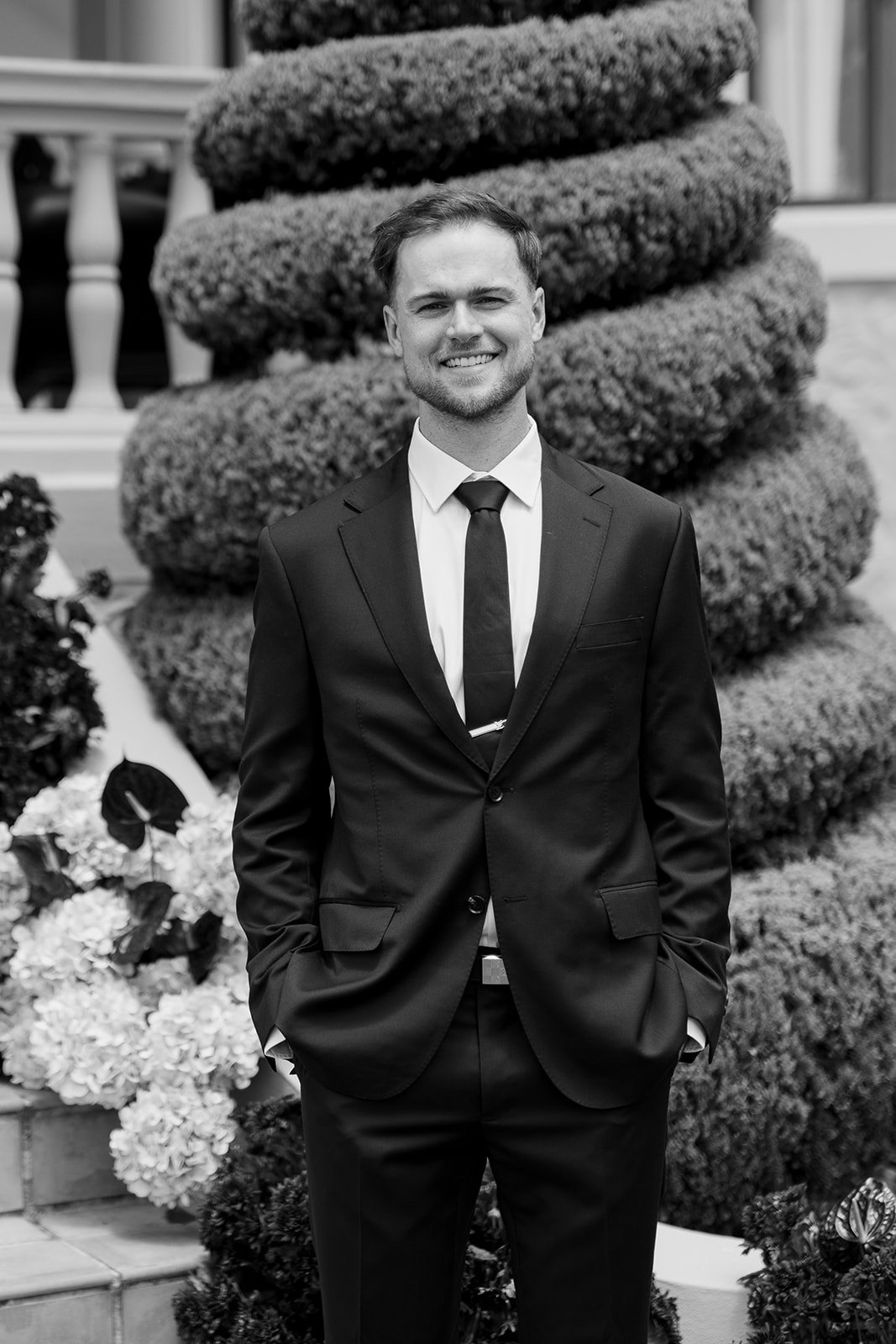 A smiling man in a suit and tie standing outdoors in front of decorative bushes and flowers.