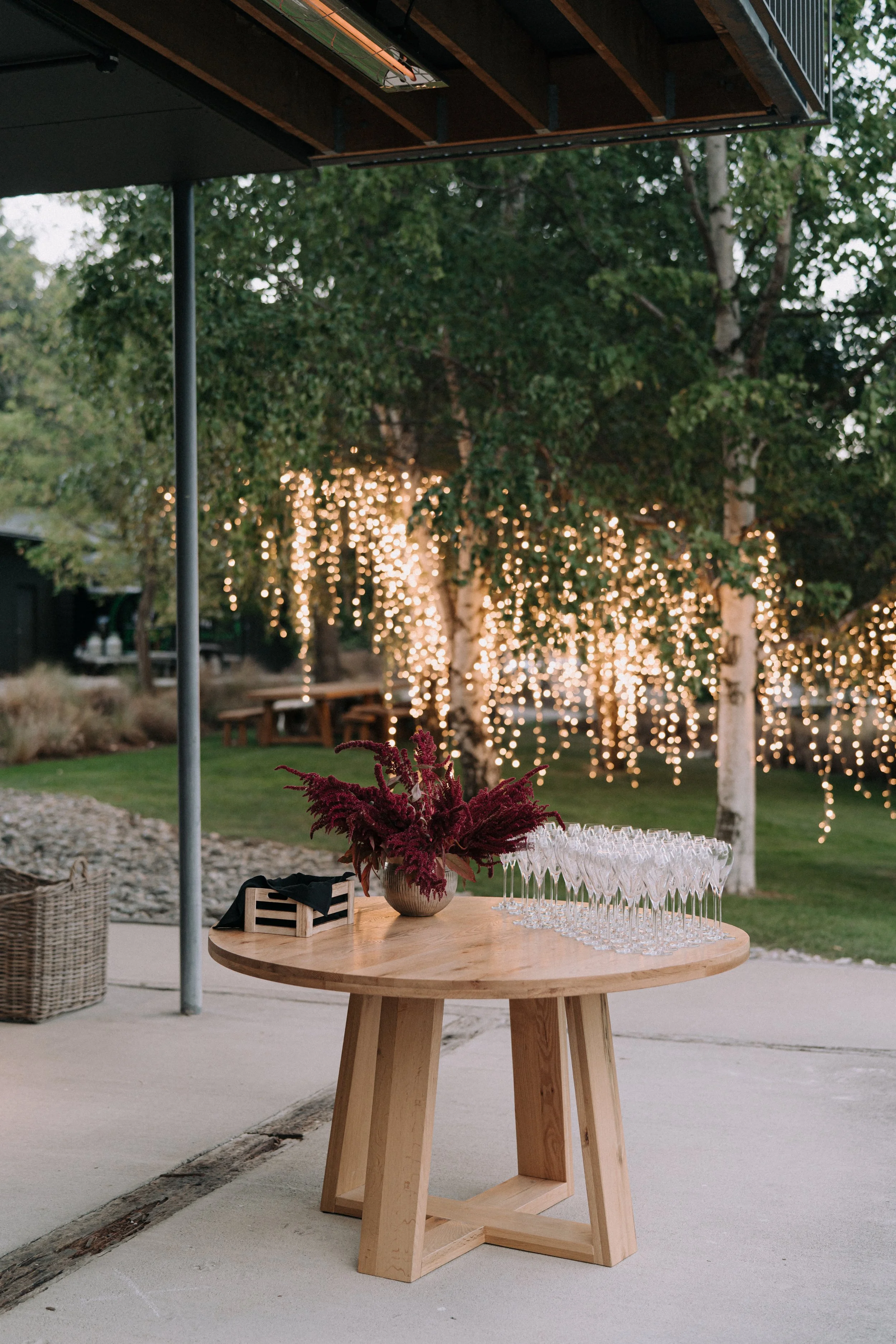 Outdoor event setup with a wooden round table holding a potted plant with burgundy flowers and several empty wine glasses, surrounded by trees with string lights, and patio furniture in the background.