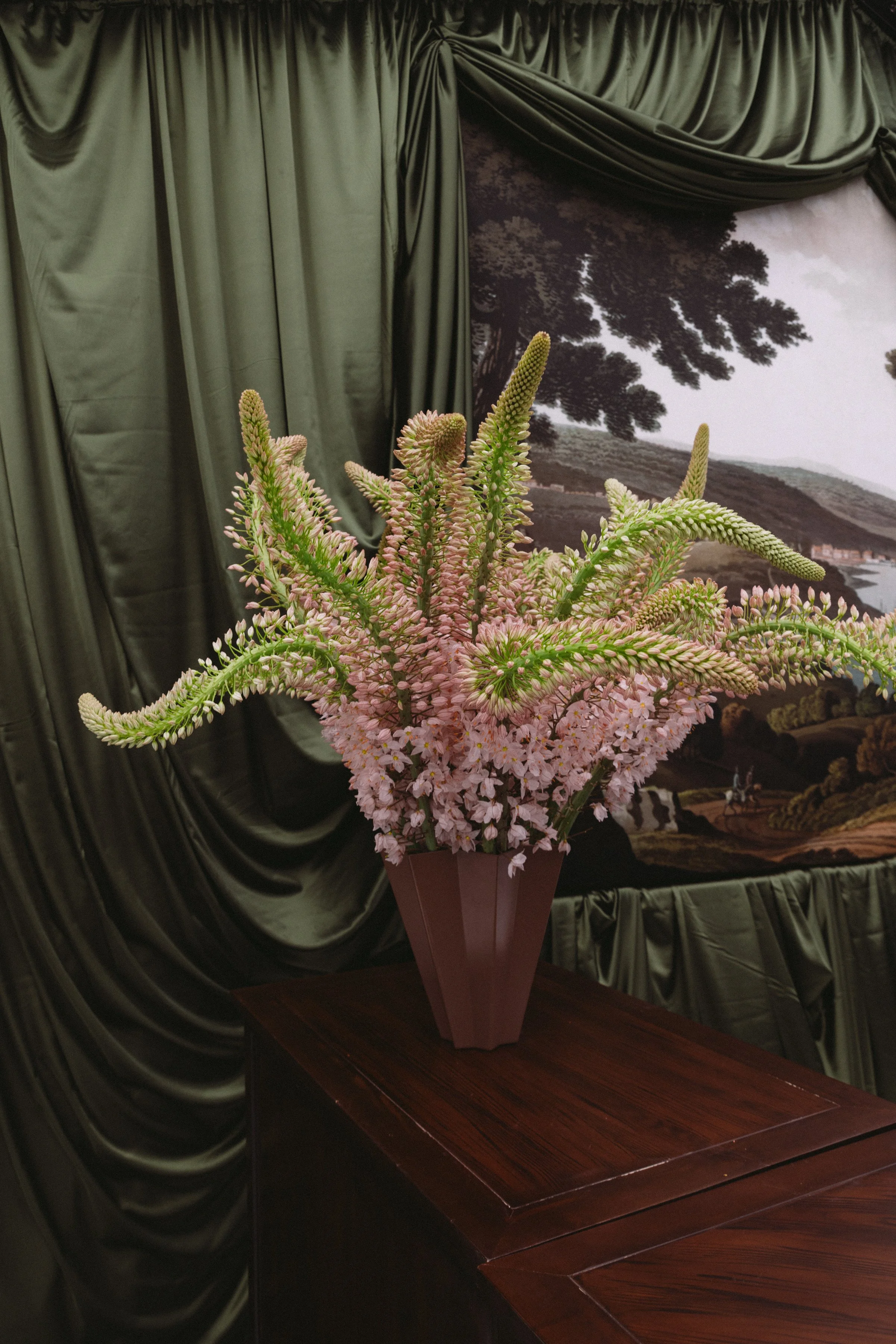 A large bouquet of pink and cream flowers in a brown vase on a wooden table, with dark green curtains and a landscape mural in the background.