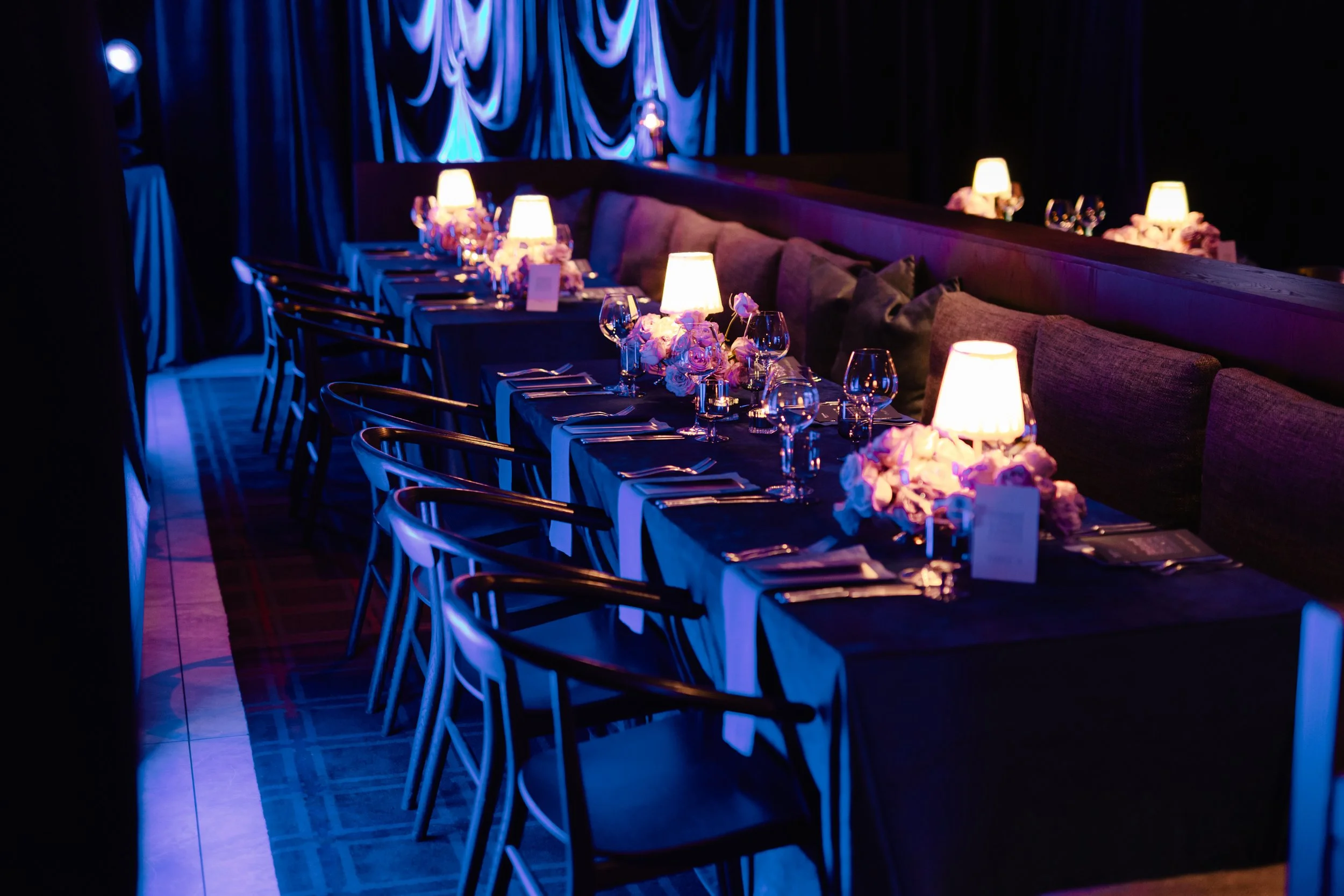 A dimly lit restaurant or event space with a long table decorated with white lamps and flowers. The table is set with glassware and utensils, and there are chairs along the sides. Blue curtains with folds are visible in the background.
