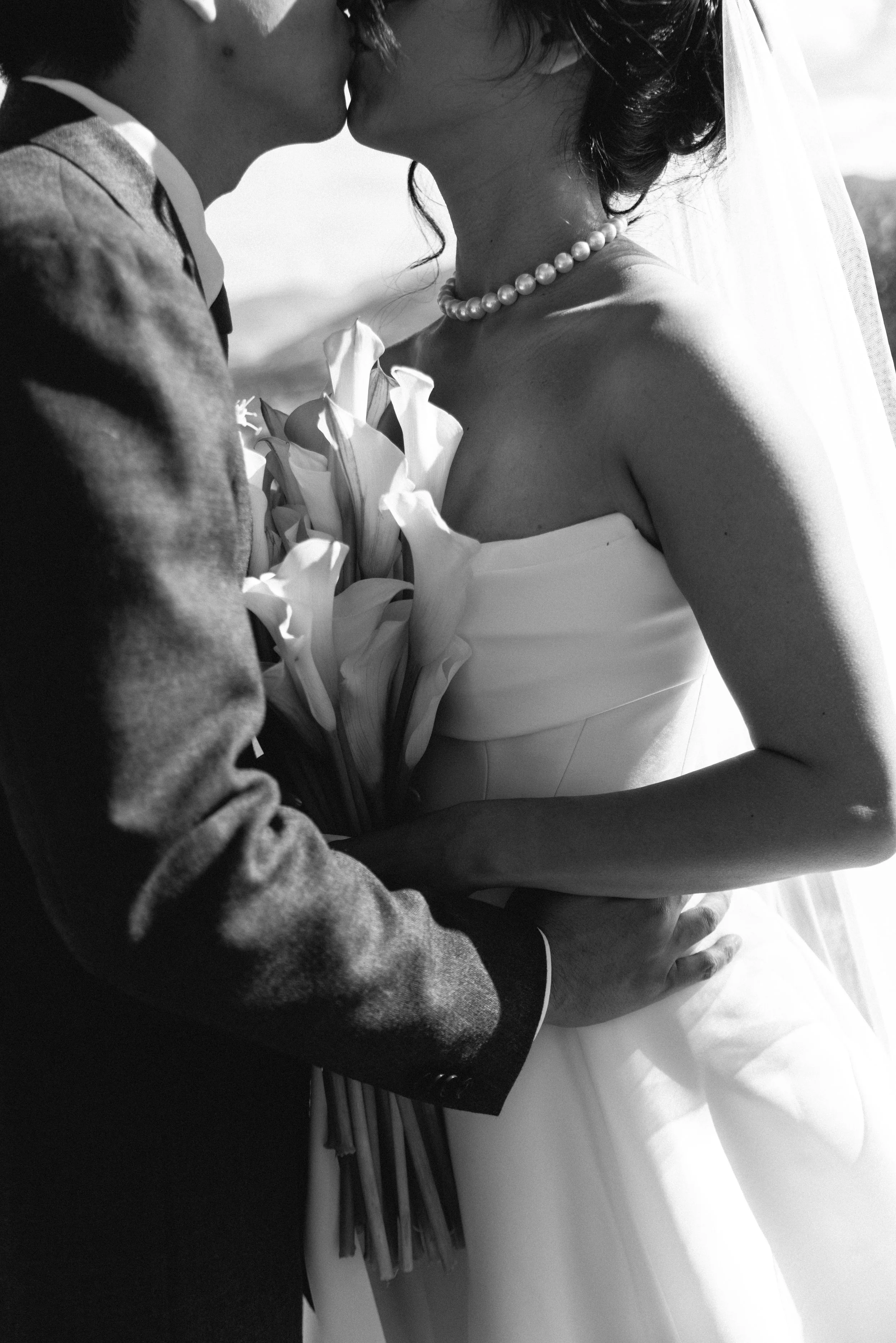A black and white photo of a couple kissing, with the man in a suit and the woman wearing a wedding dress and pearl necklace.