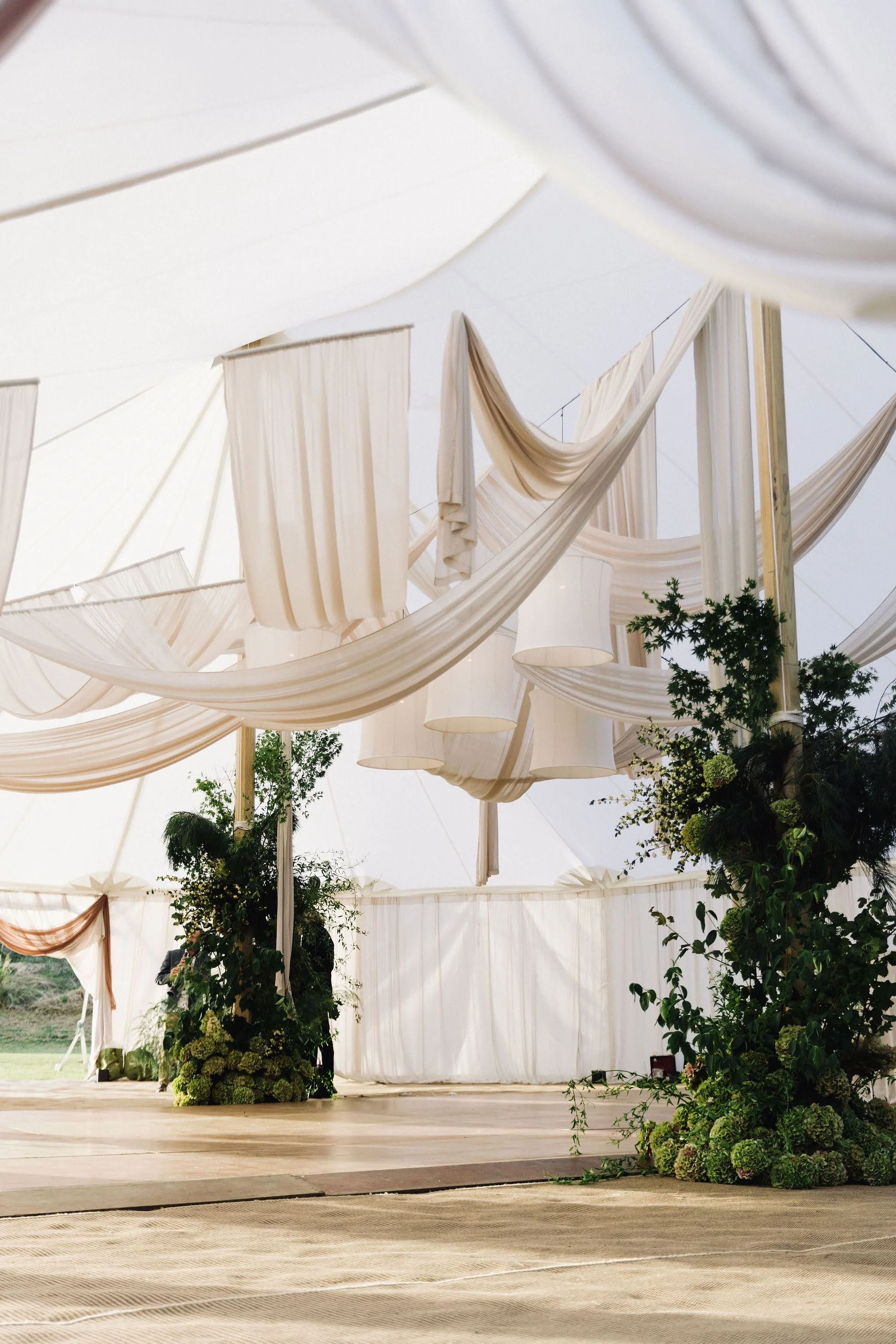 A white event tent with draped fabric, hanging lamps, and greenery decorations.