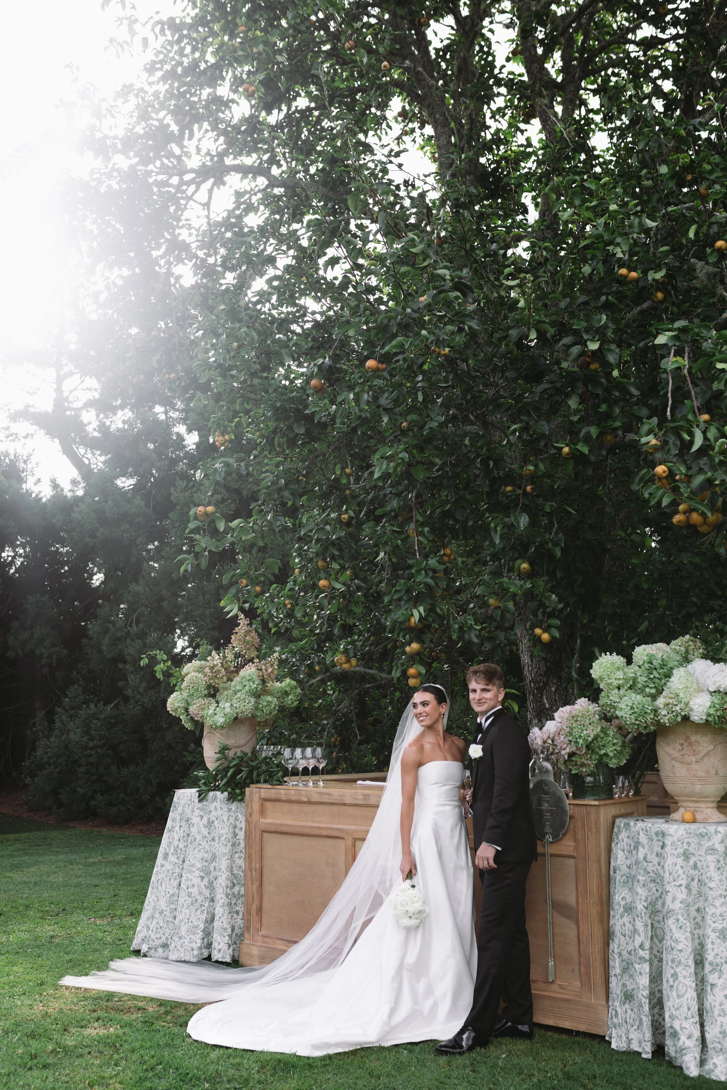 A bride and groom stand outdoors during a wedding reception. The bride wears a white strapless wedding gown with a long train and veil, holding a bouquet of white flowers. The groom wears a black tuxedo with a white boutonniere. They are smiling in front of a wooden bar decorated with large vases of white and pale pink hydrangeas, lemons, and greenery, set in a garden with trees and greenery in the background.