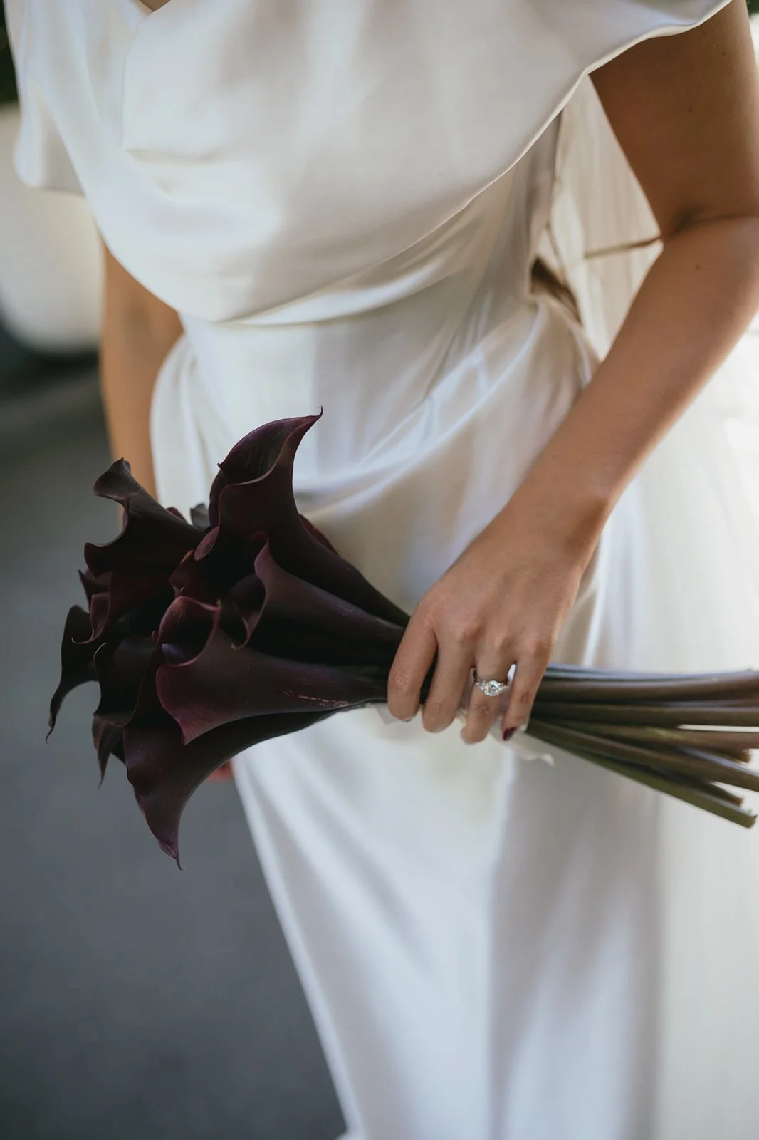 A woman in a white dress holding a dark purple calla lily bouquet with her left hand, showing a diamond ring on her ring finger.