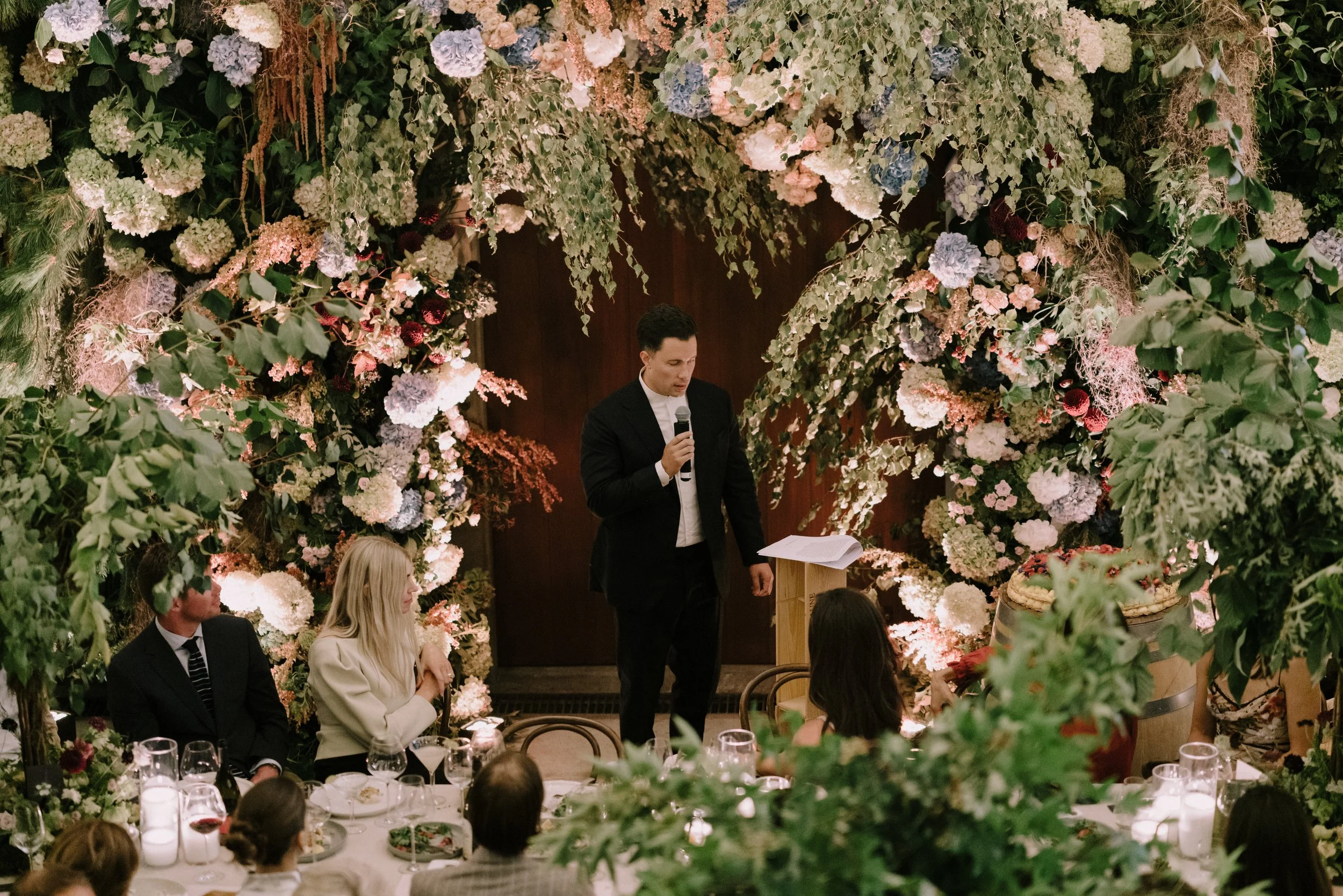 A man in a tuxedo speaking into a microphone during a formal event, with a floral arch made of various flowers and greenery overhead and around him. Guests are seated at decorated tables in an elegant setting.