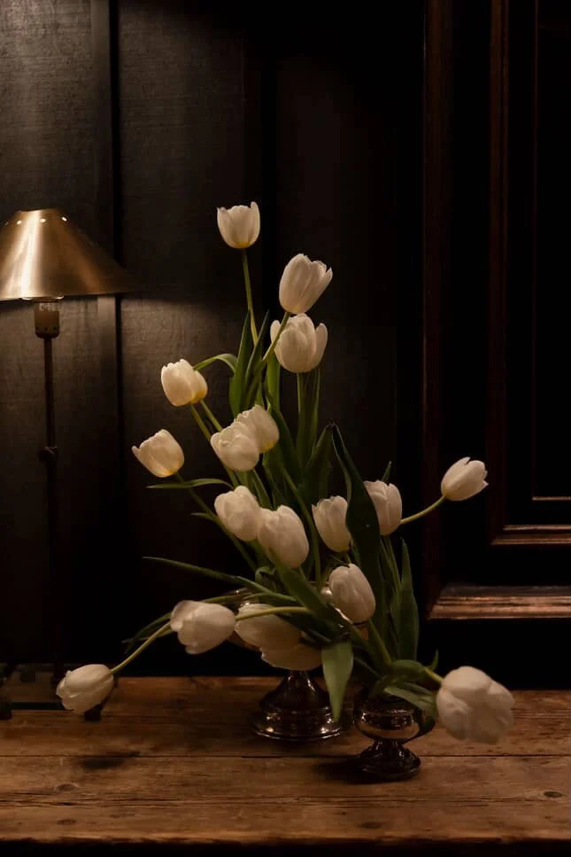 A bouquet of white tulips in a dark-colored vase on a wooden surface, with a lamp in the background and dark walls.