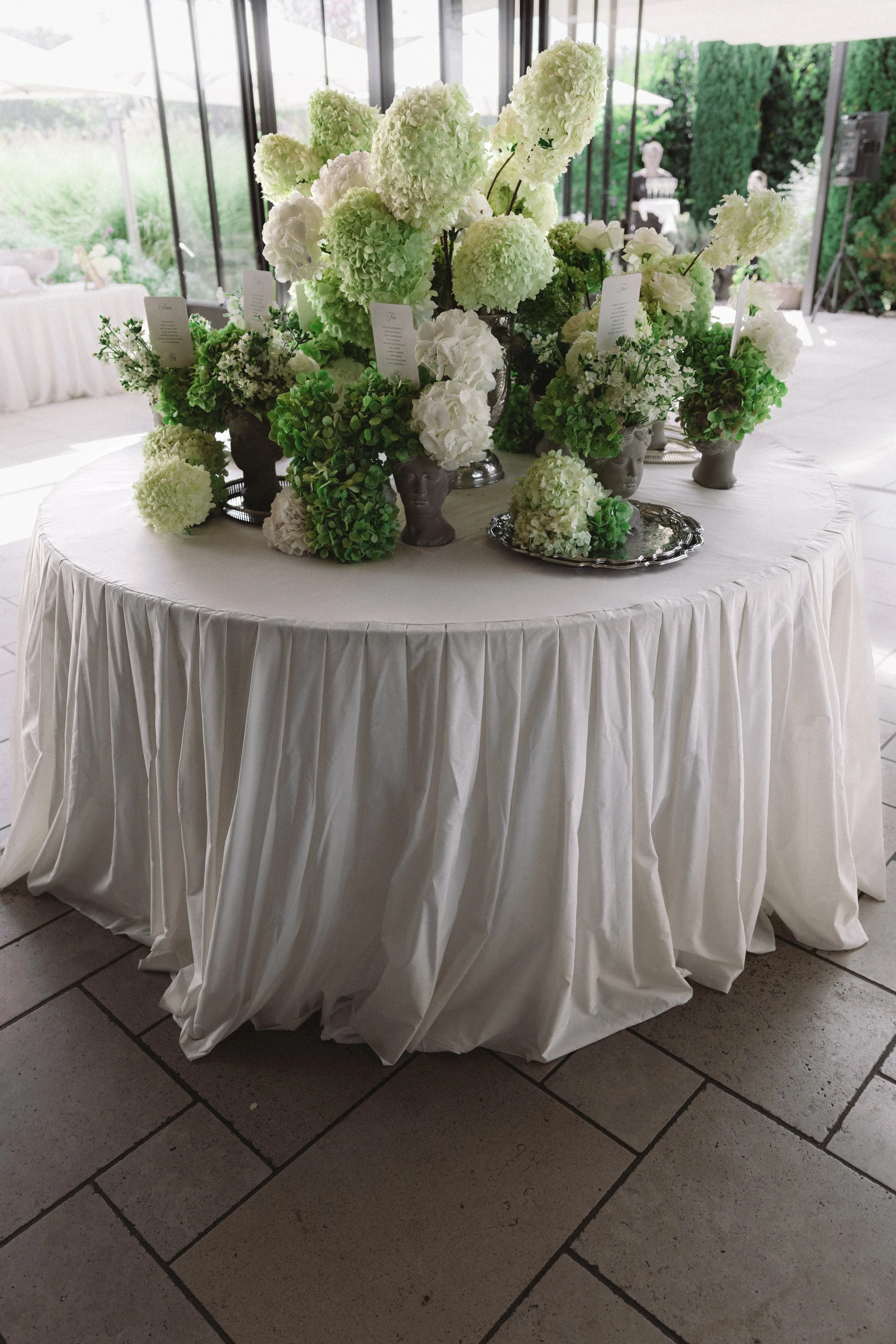 A round table draped with a white tablecloth with pleats, holding a large floral centerpiece of white hydrangeas and other white flowers in various vases. Small cards are inserted into the floral arrangement. The background features large windows and a garden with green shrubs and outdoor furniture.