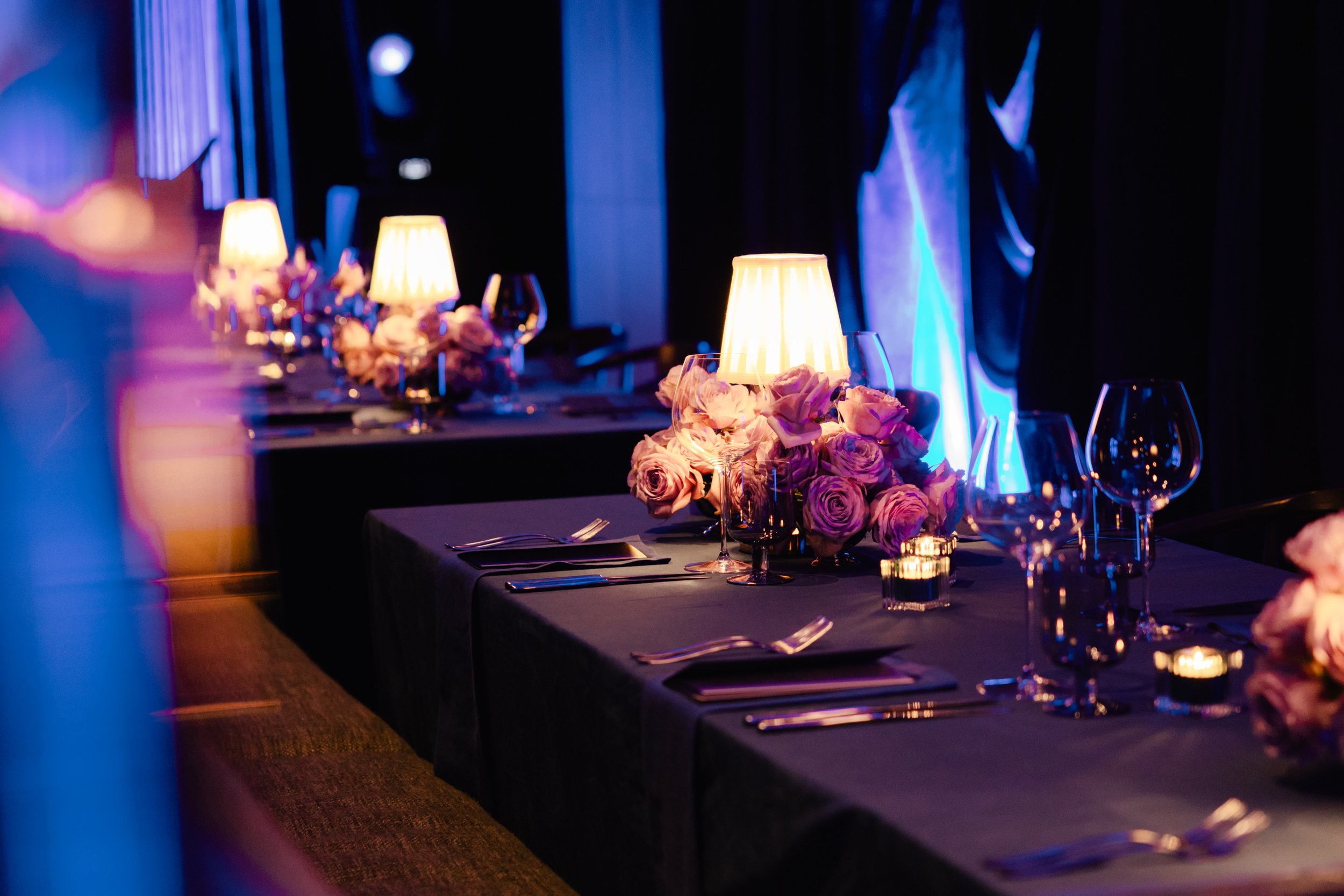 A long dining table decorated with pink roses, wine glasses, candles, and tableware, set for a formal event in a dimly lit room with blue accent lighting.