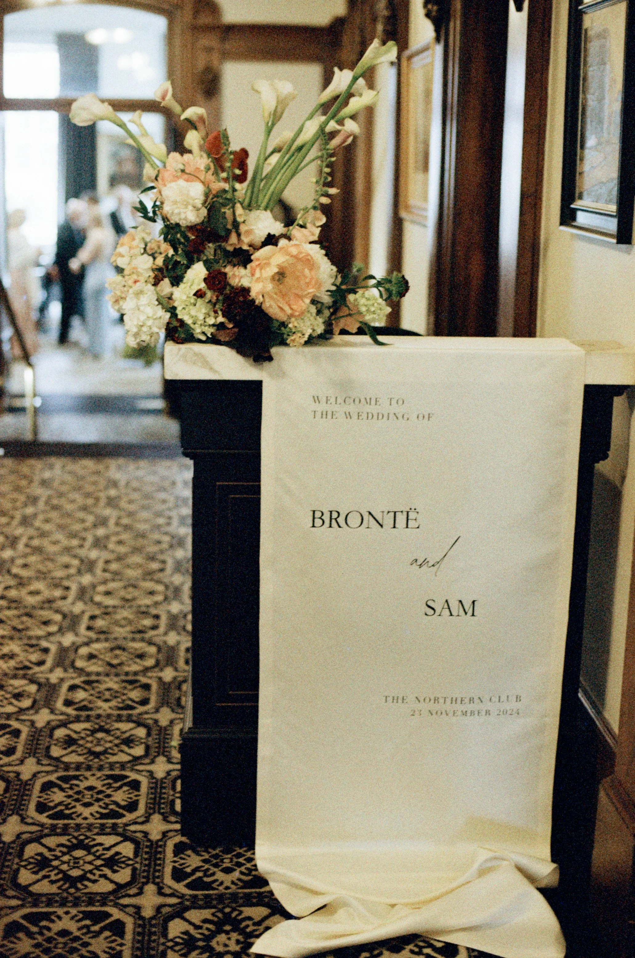 A floral arrangement on a wedding welcome sign at a reception, with blurred people in the background.