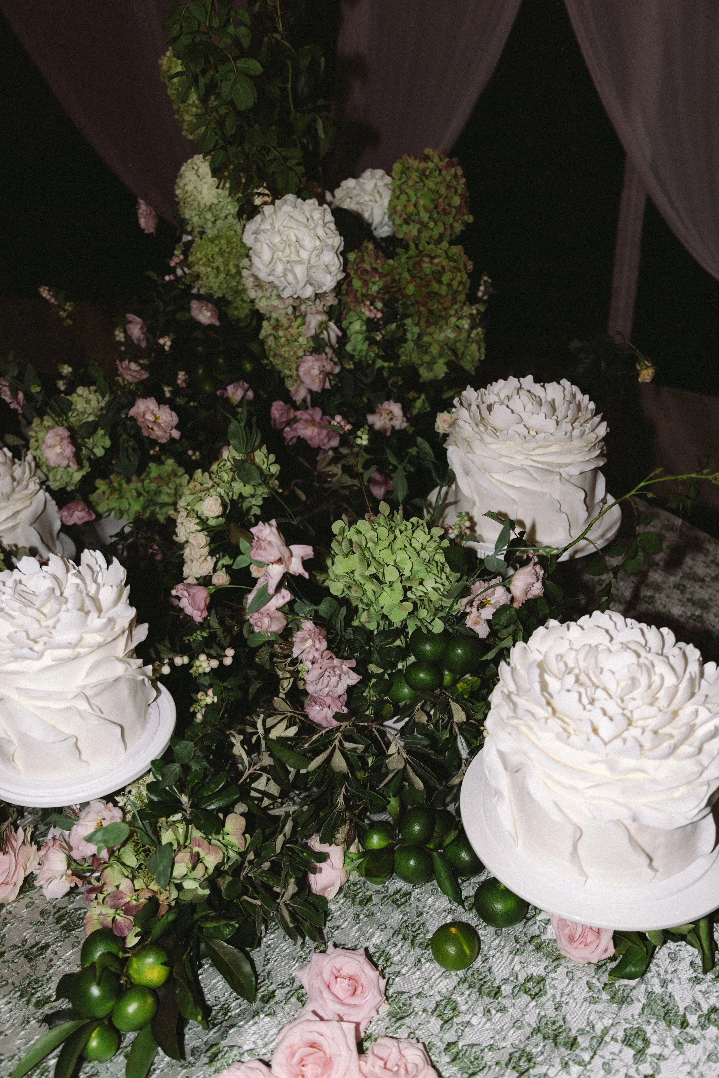 Close-up of a floral arrangement with white roses, green and pink hydrangeas, pink roses, green berries, and green leaves on a table with a textured cloth and pink flowers scattered around.