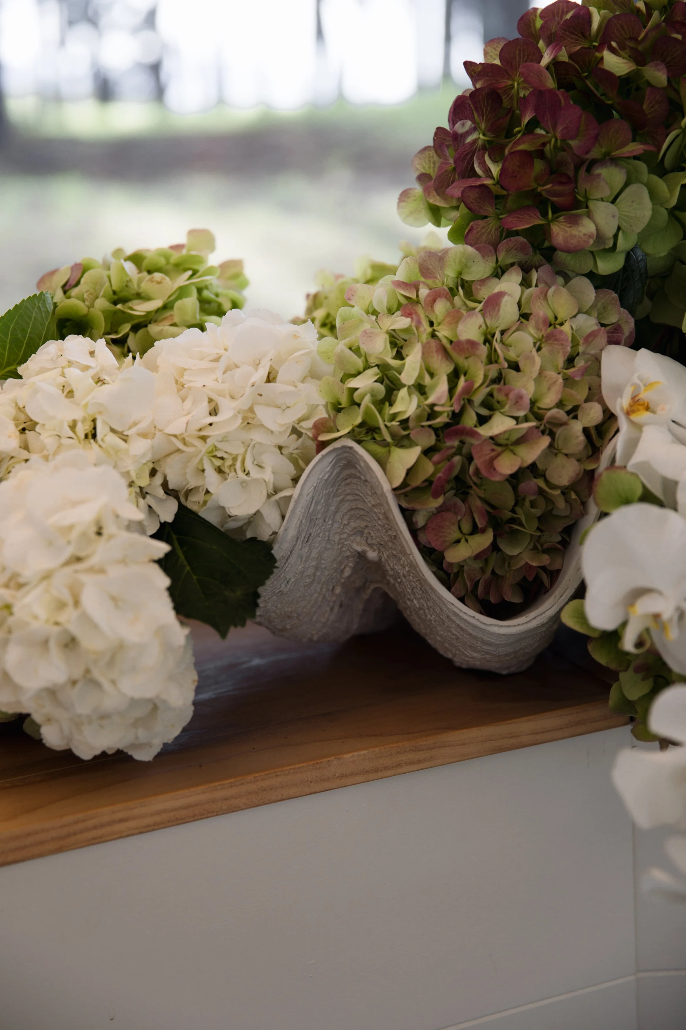 Close-up of a wooden tray with white and pinkish-green hydrangea flowers, some orchids, and a decorative piece, with a blurred background of windows and light.