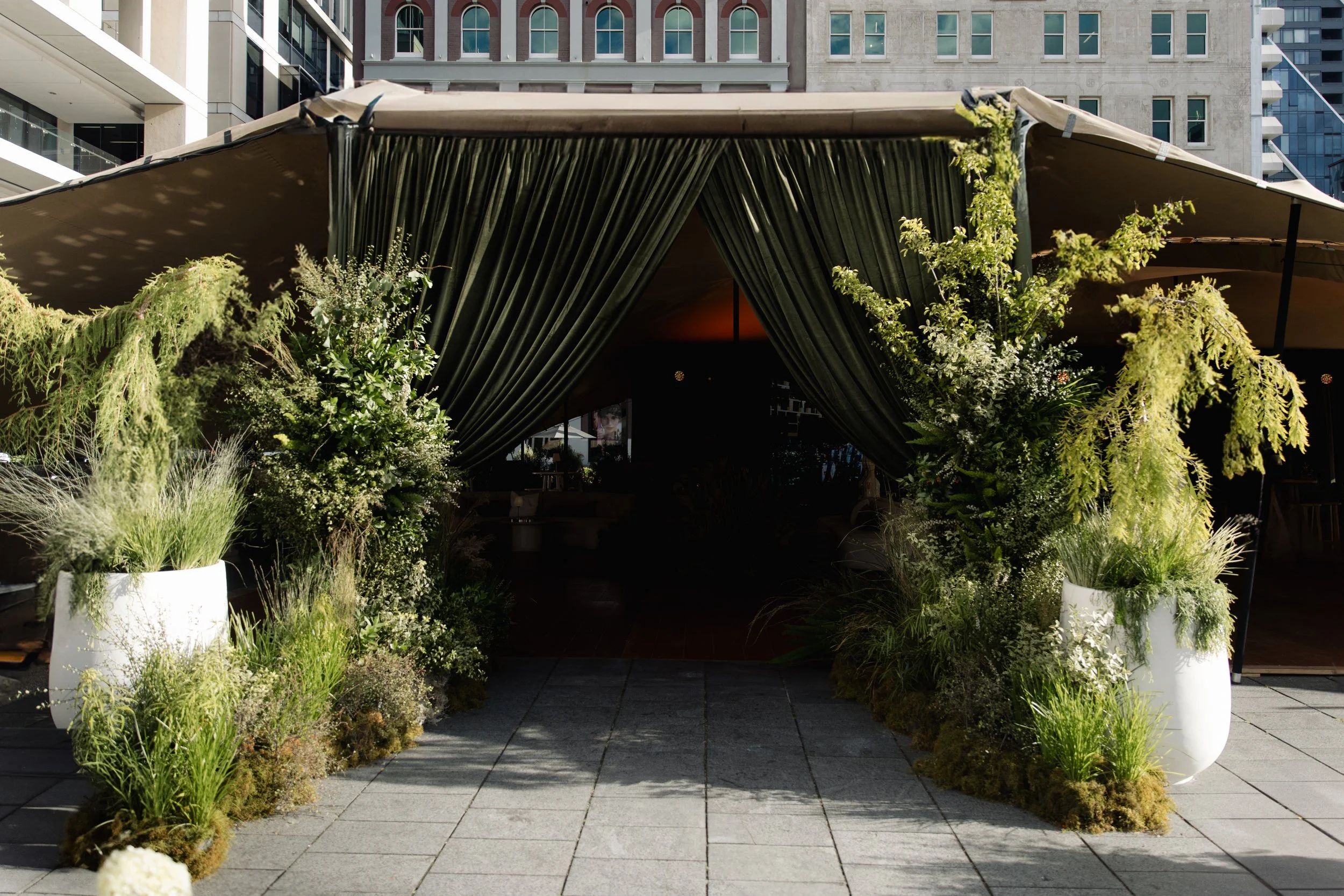 An outdoor event space with a large tent decorated with green curtains and surrounded by green plants and white flower arrangements, set against tall buildings in an urban area.
