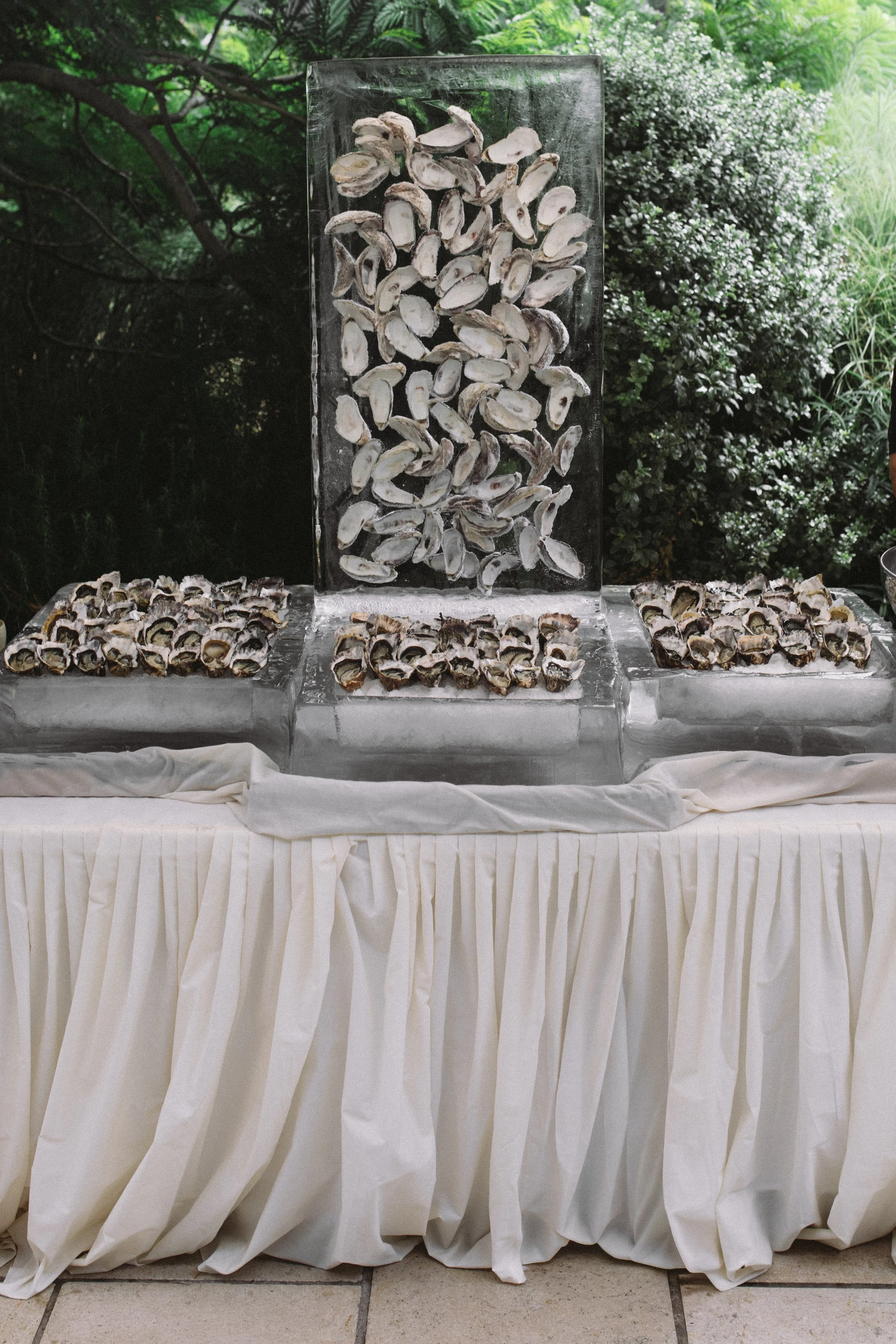 A table with a white cloth skirt, topped with ice blocks holding oyster shells, set outdoors with dense green foliage in the background.
