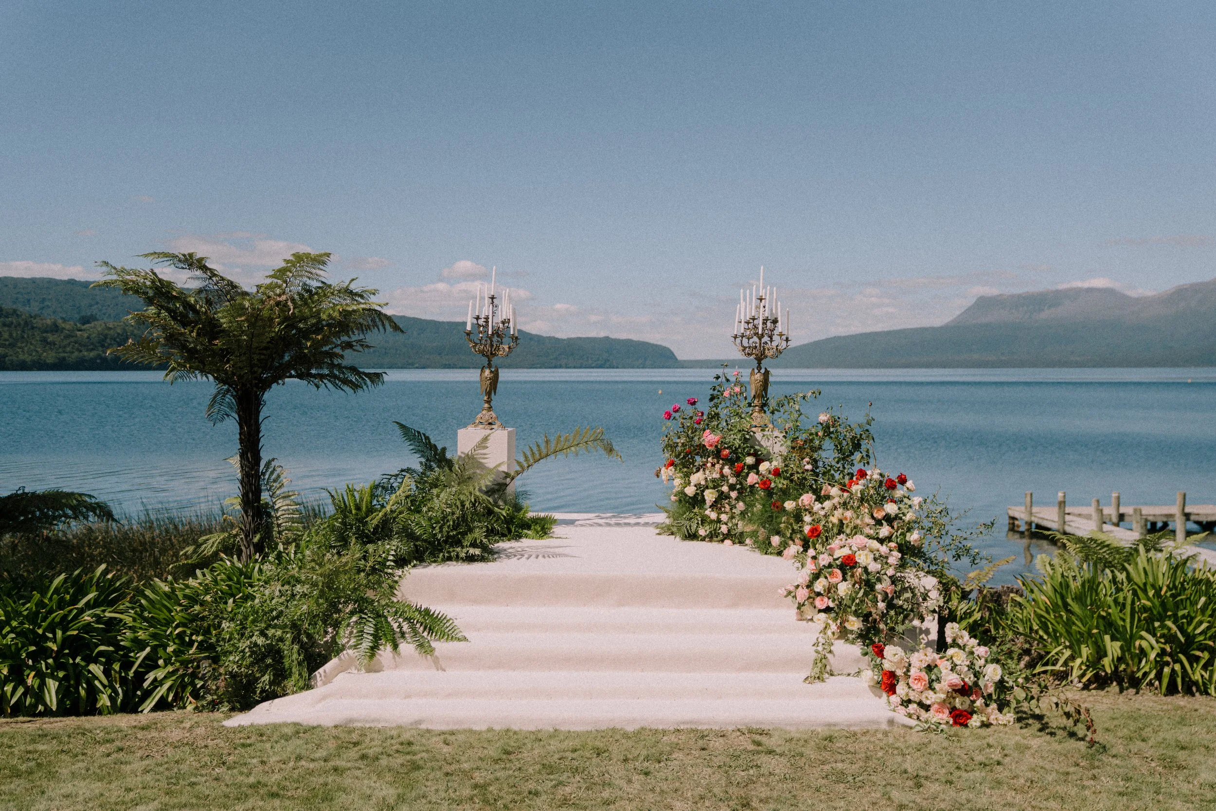 A lakeside scene with a platform decorated with chandeliers, flowers, and greenery, overlooking a calm lake with mountains in the background.
