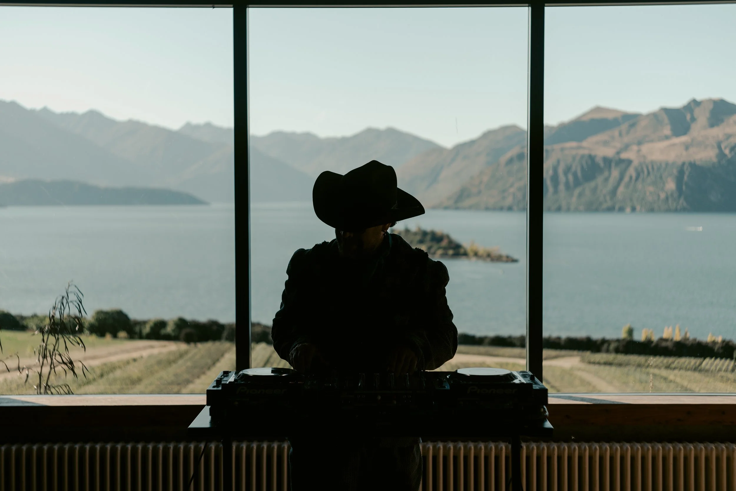 Silhouette of a DJ wearing a cowboy hat, standing behind a DJ mixer, in front of large windows overlooking mountains and a lake.