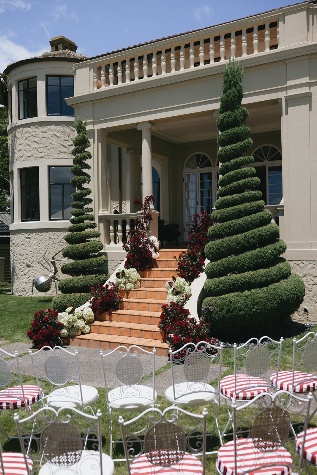 A large, elegant house with a entry staircase surrounded by manicured topiary trees and decorative flowers, set up for an outdoor event with white chairs that have red and white striped cushions.