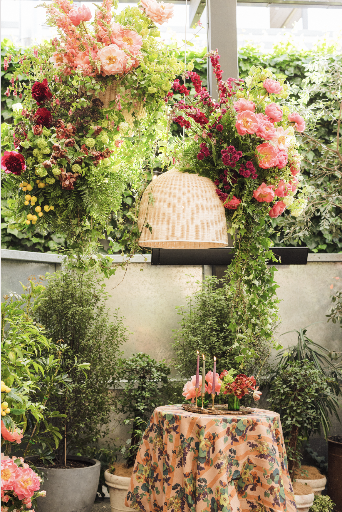A lush garden scene with abundant pink, red, and green flowers and foliage. A table with a floral tablecloth and candles is in the foreground, and hanging flower arrangements are above, creating a vibrant and decorative atmosphere.