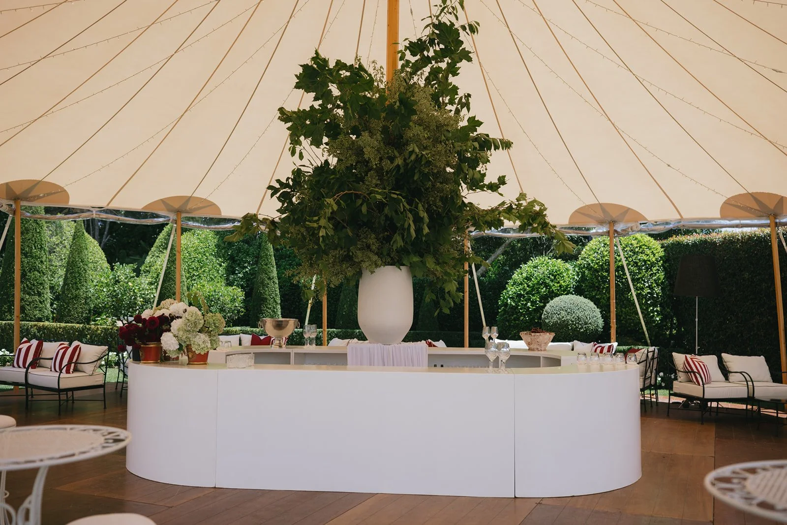 A large floral arrangement in a white vase on an outdoor party or event table with a white draped fabric covering the front, under a tent with string lights. The background features lush green trees and shrubs, with seating areas including white cushioned chairs with striped pillows.