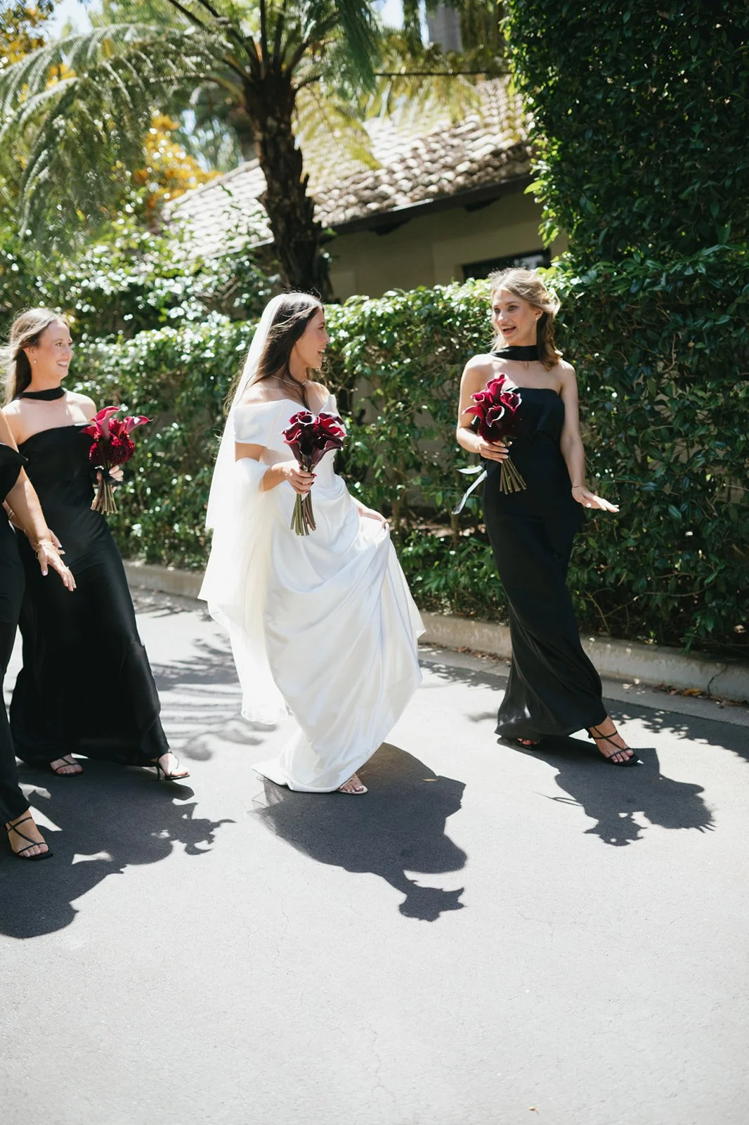 A group of women walking outdoors on a sunny day, with one woman wearing a white wedding dress and veil, holding a bouquet of dark red calla lilies, and the other women in black dresses also holding bouquets, smiling and talking to each other.