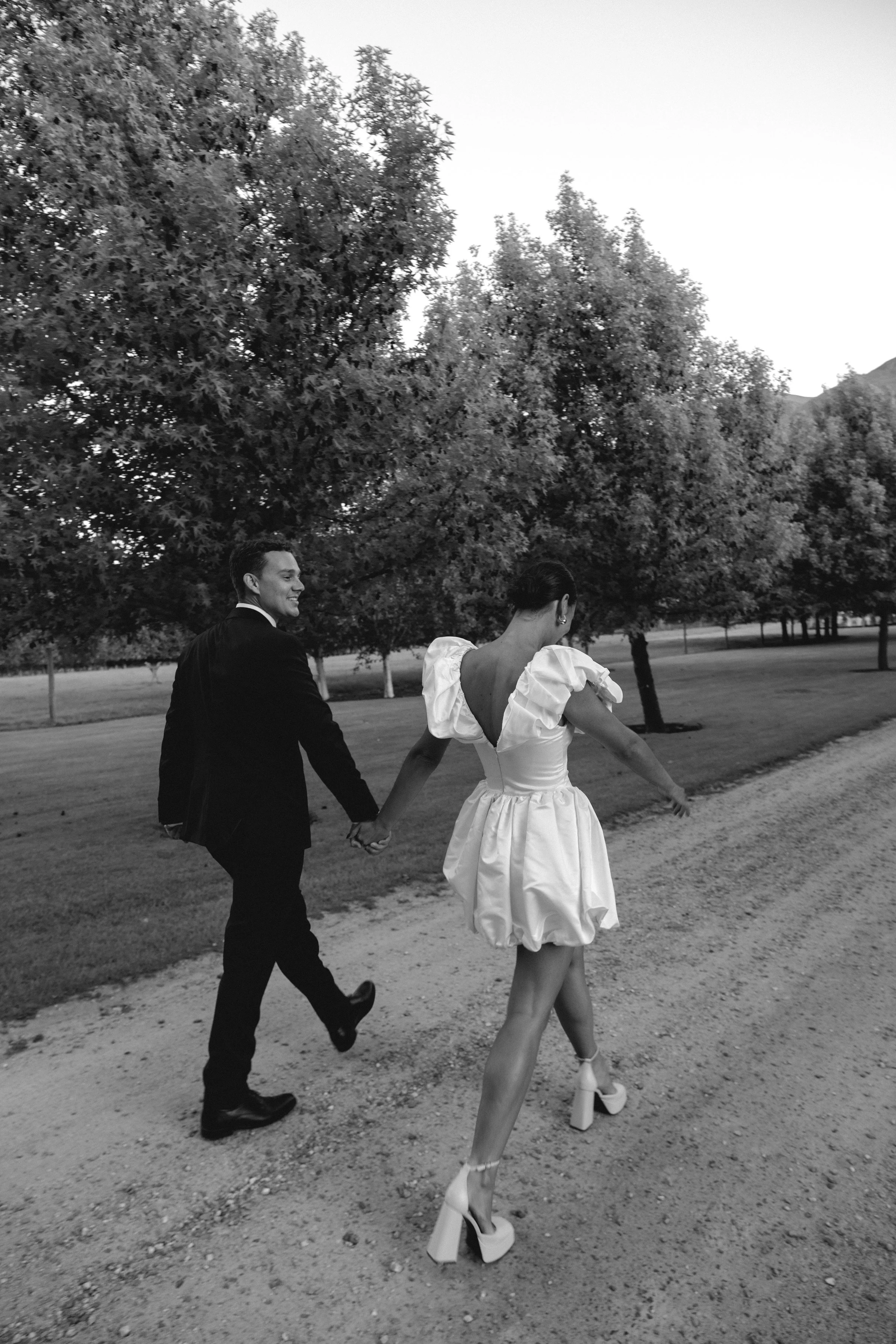 A black and white photo of a couple holding hands and walking outdoors in a park with trees. The woman is wearing a stylish, backless dress with exaggerated puffy sleeves and high platform heels. The man is in a dark suit. They are smiling and looking at each other, enjoying a moment together.