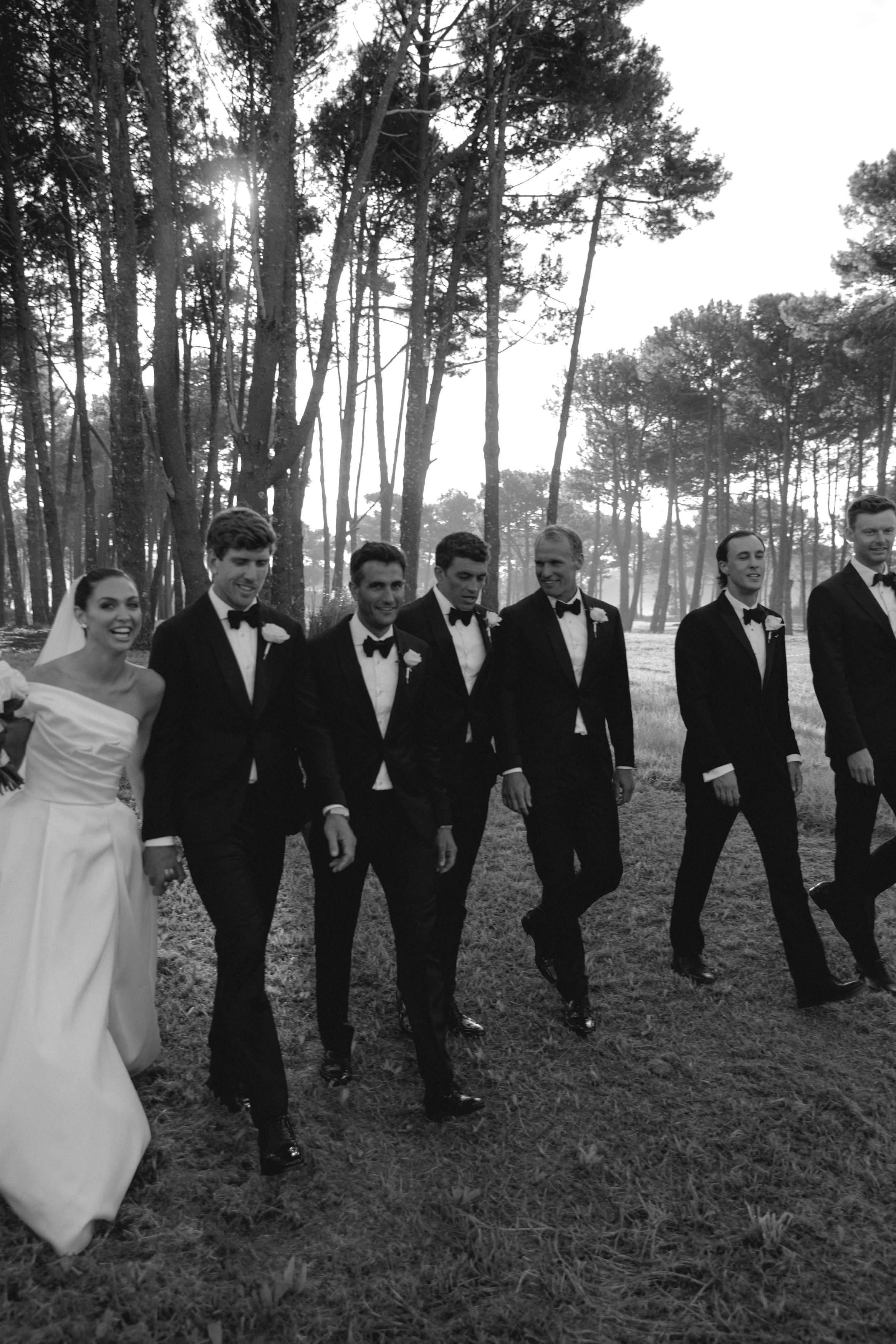 A black and white photo of a wedding party walking outdoors through a grassy area surrounded by tall trees, with the sun shining through the trees.