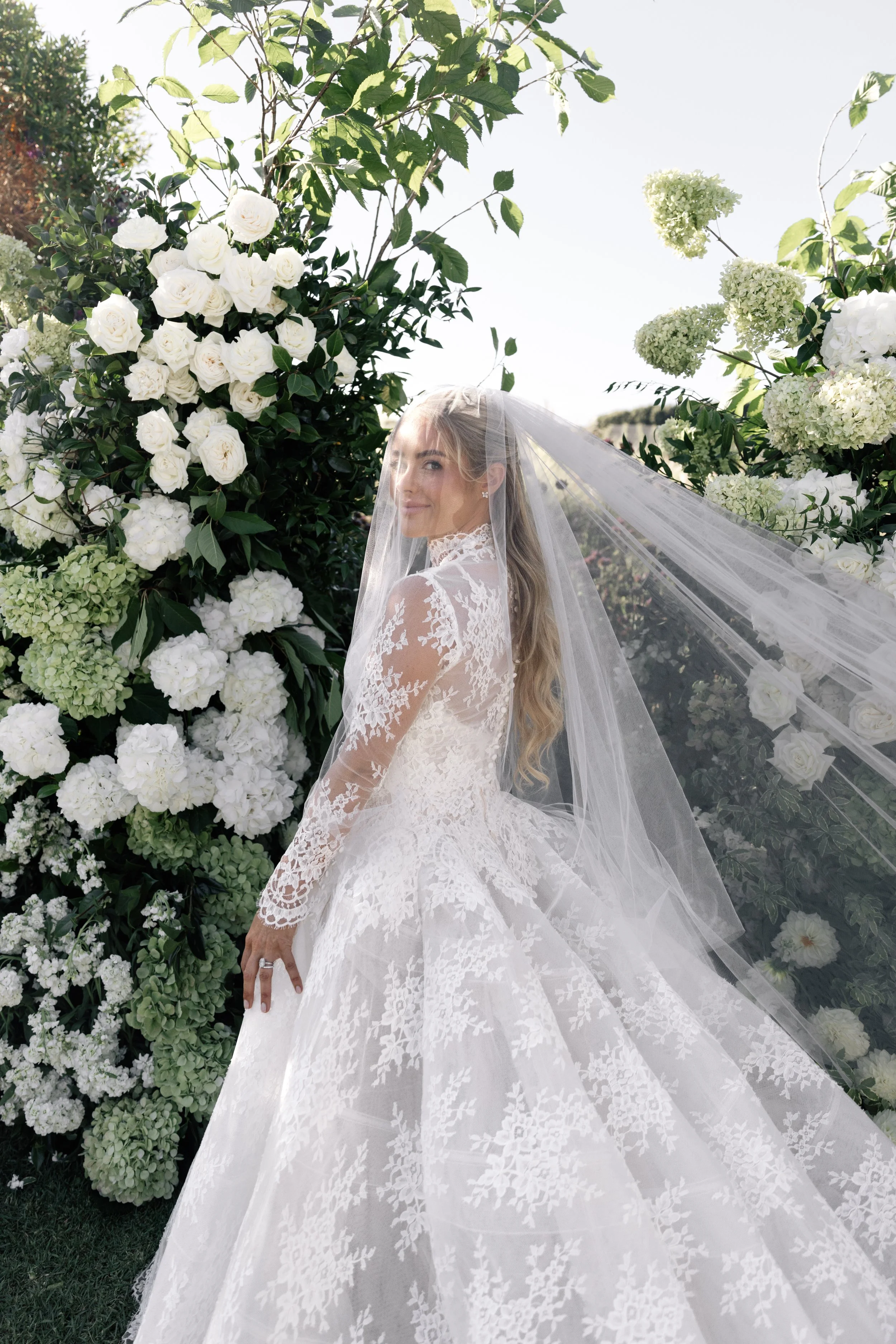 A bride in a white lace wedding gown with long sleeves and a veil, standing amidst lush white flowers and greenery outdoors during the day.