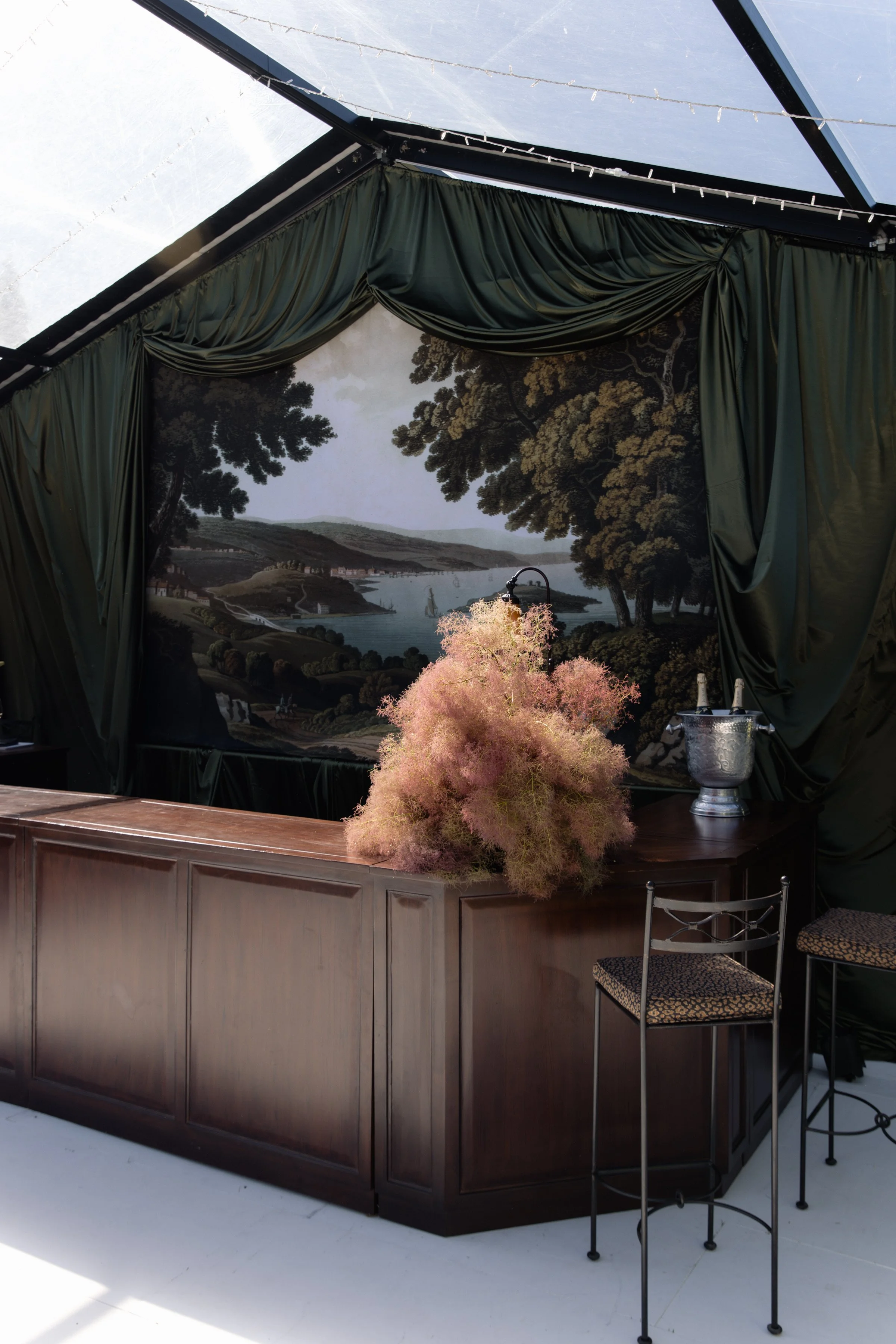 An indoor bar with dark wood paneling and a painted landscape backdrop of trees, a river, and hills. Green curtains hang above, and a potted pink plant sits on the bar counter. Two metal chairs with patterned cushions are in front of the bar, and a small table with a bottle and a glass is nearby.