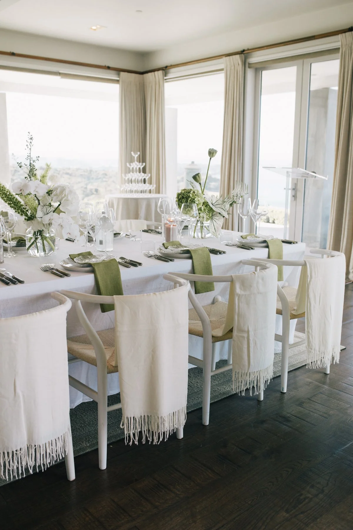 Elegant banquet table setup with white tablecloth, floral centerpieces, wine glasses, and green napkins, in a bright room with large windows and curtains.