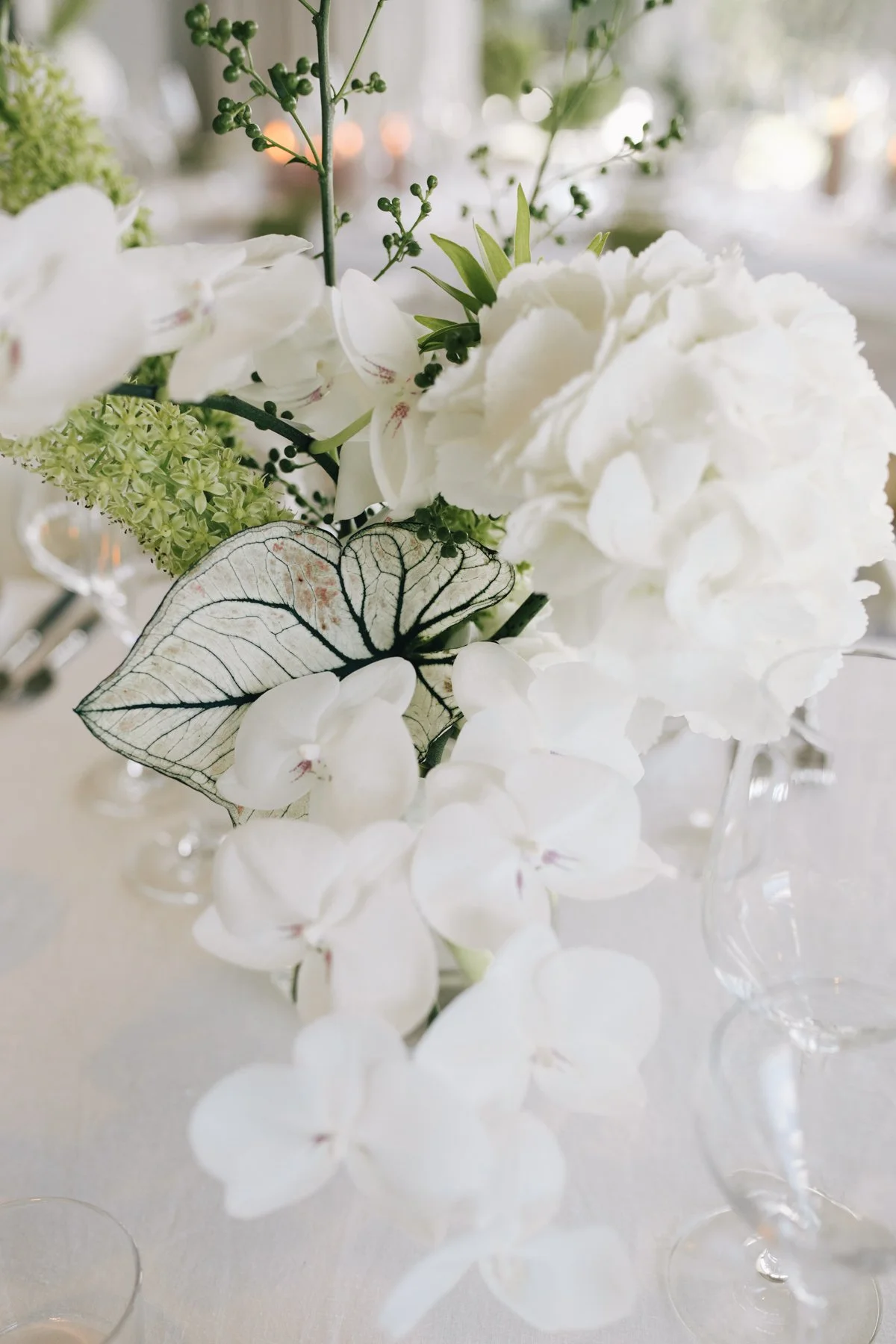 Close-up of a white floral arrangement featuring hydrangeas, orchids, greenery, and a leaf with visible veins, displayed in a glass vase on a table with a blurred background.