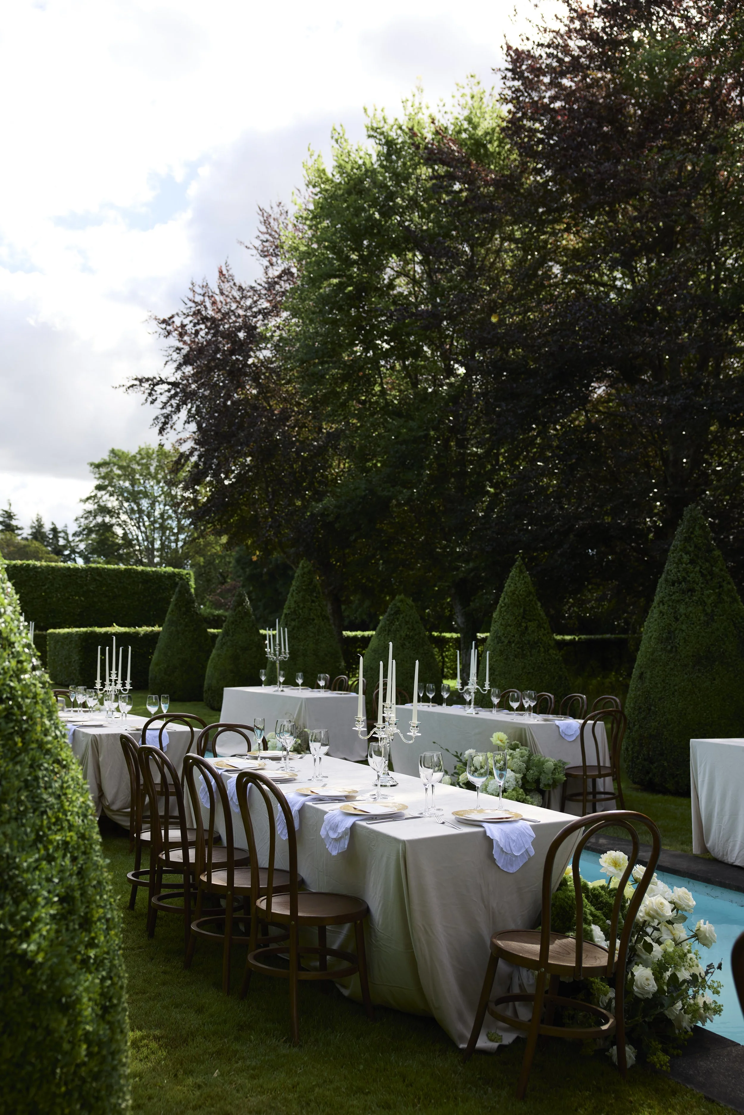 Outdoor banquet setting with long tables covered in white tablecloths, elegant glassware, candelabras, and floral centerpieces, surrounded by greenery and trees.