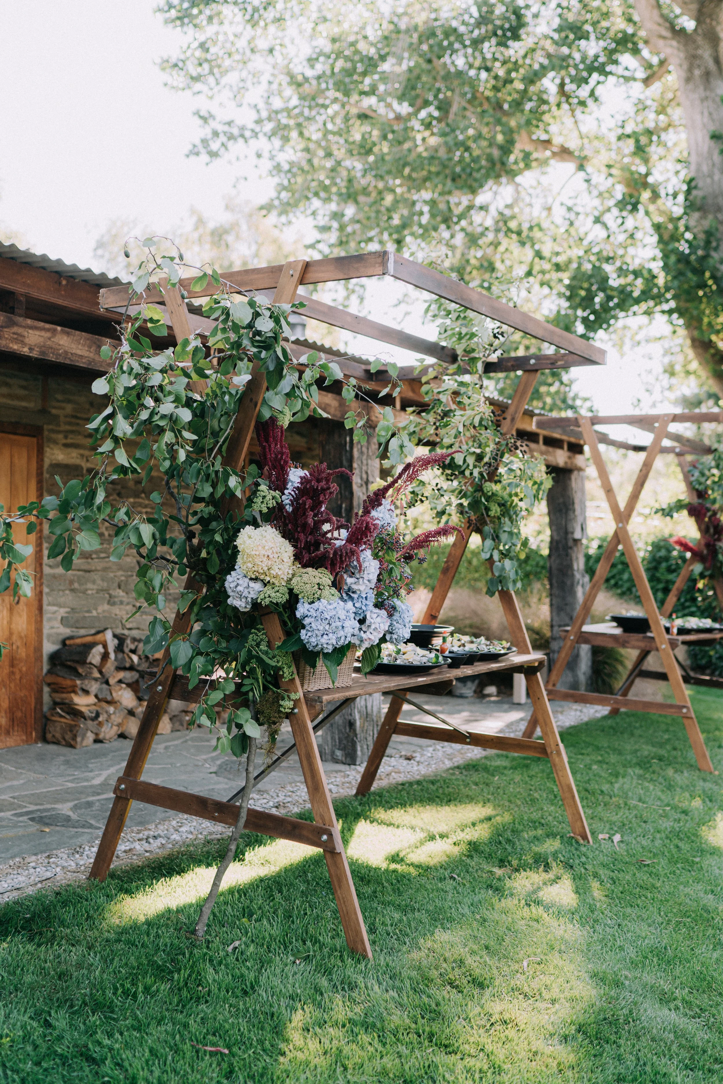 Outdoor garden setting with wooden trellises and lush greenery, including flowering plants and a large tree in the background.