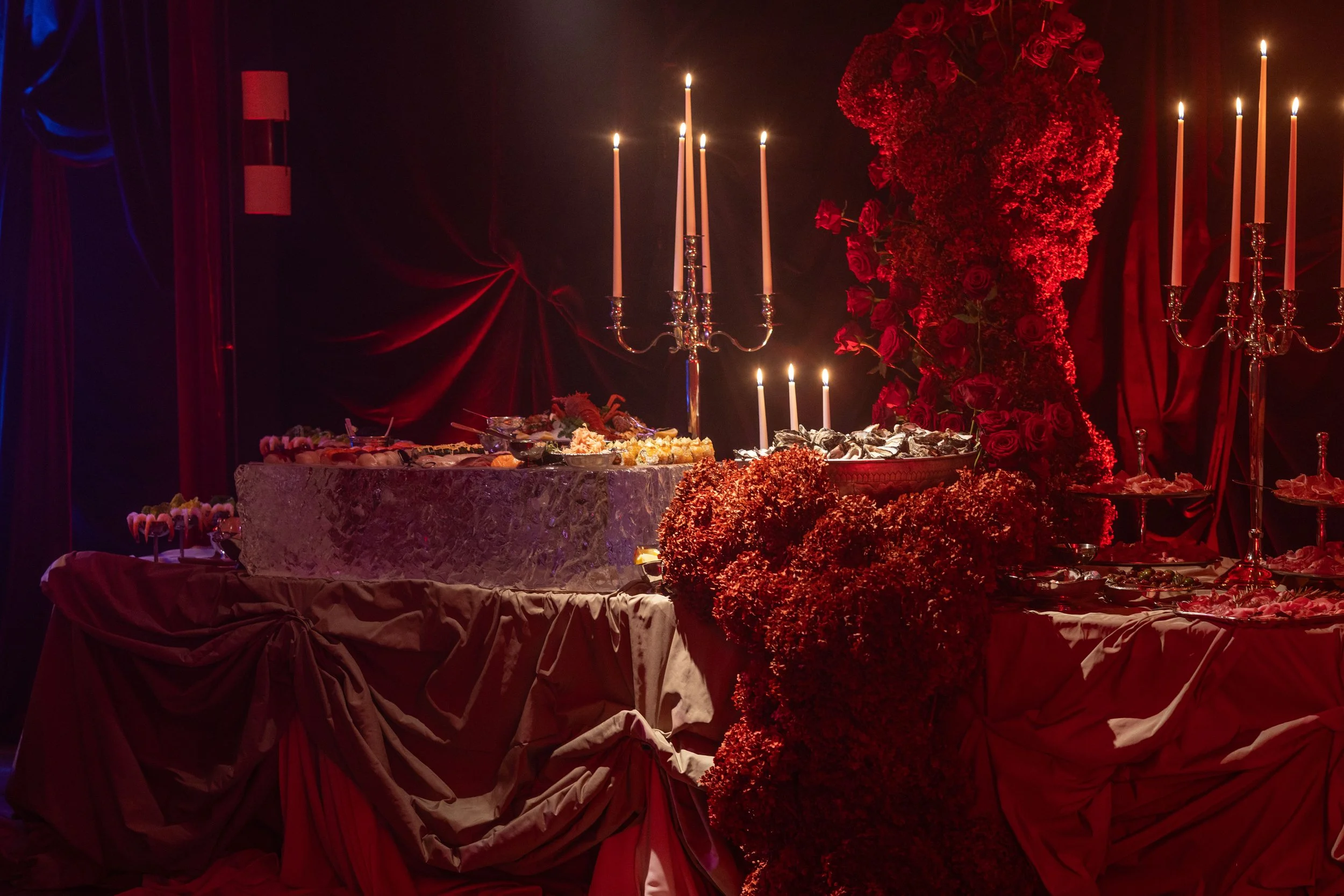 A table decorated with a large bouquet of red roses and flowers, tall lit candles in silver candleholders, and an assortment of food and desserts, set against red curtains and dim lighting, creating an elegant and romantic atmosphere.
