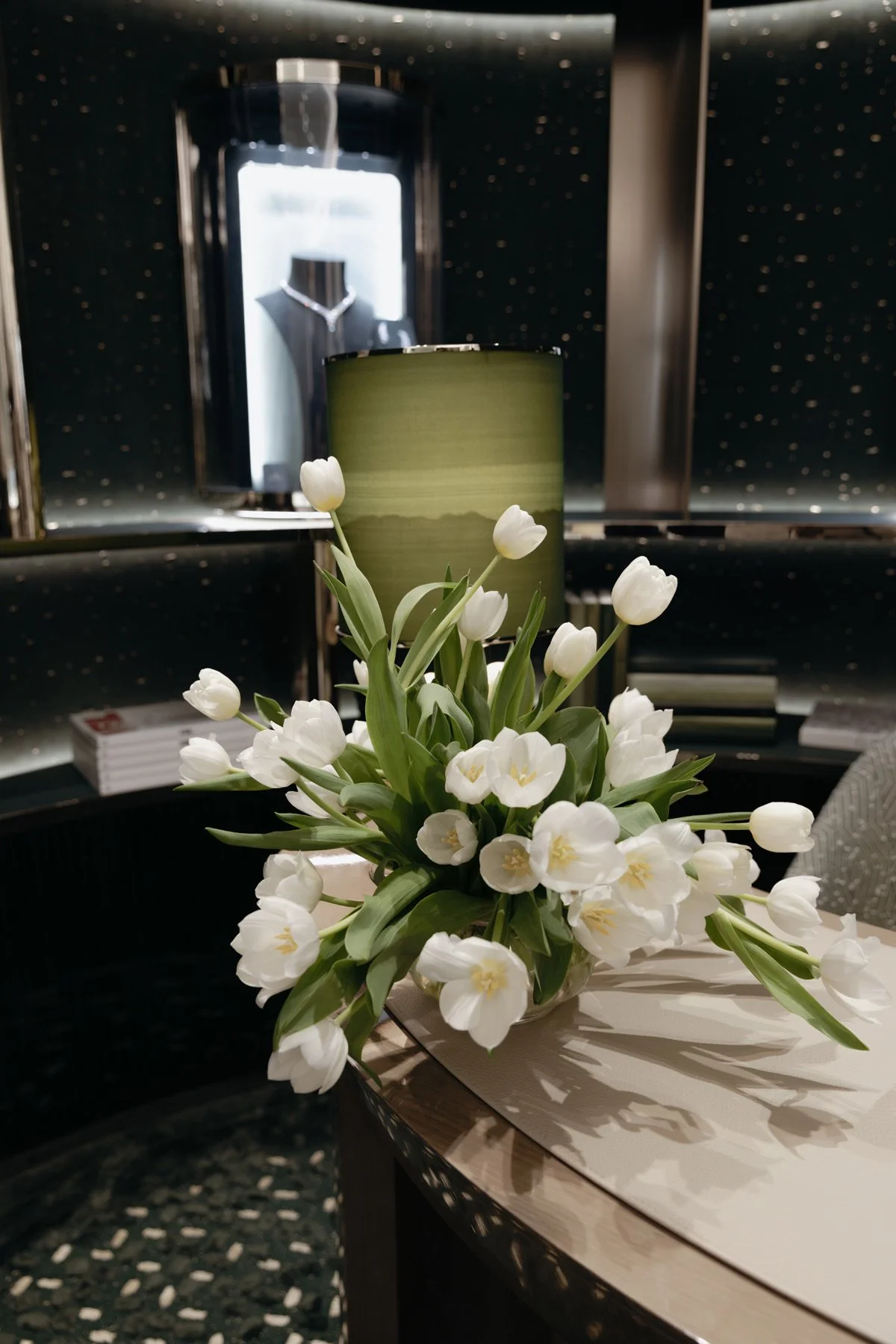 A vase of white tulips on a table with a green lampshade and jewelry display in the background.