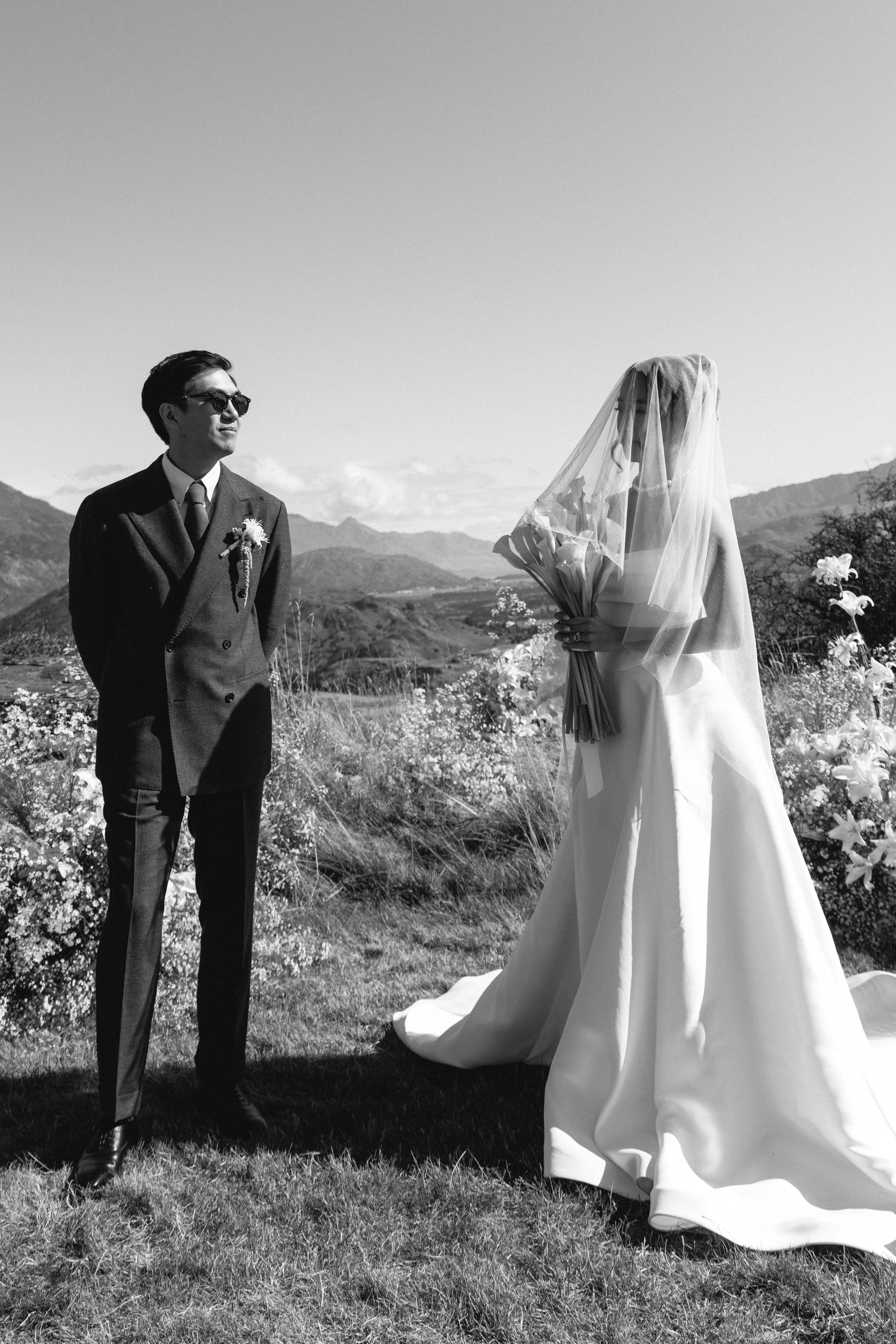 A black-and-white photo of a bride with a veil holding a bouquet of flowers on her wedding day, standing outdoors on grass with mountains in the background. A groom in a suit wearing sunglasses stands nearby, looking at her.