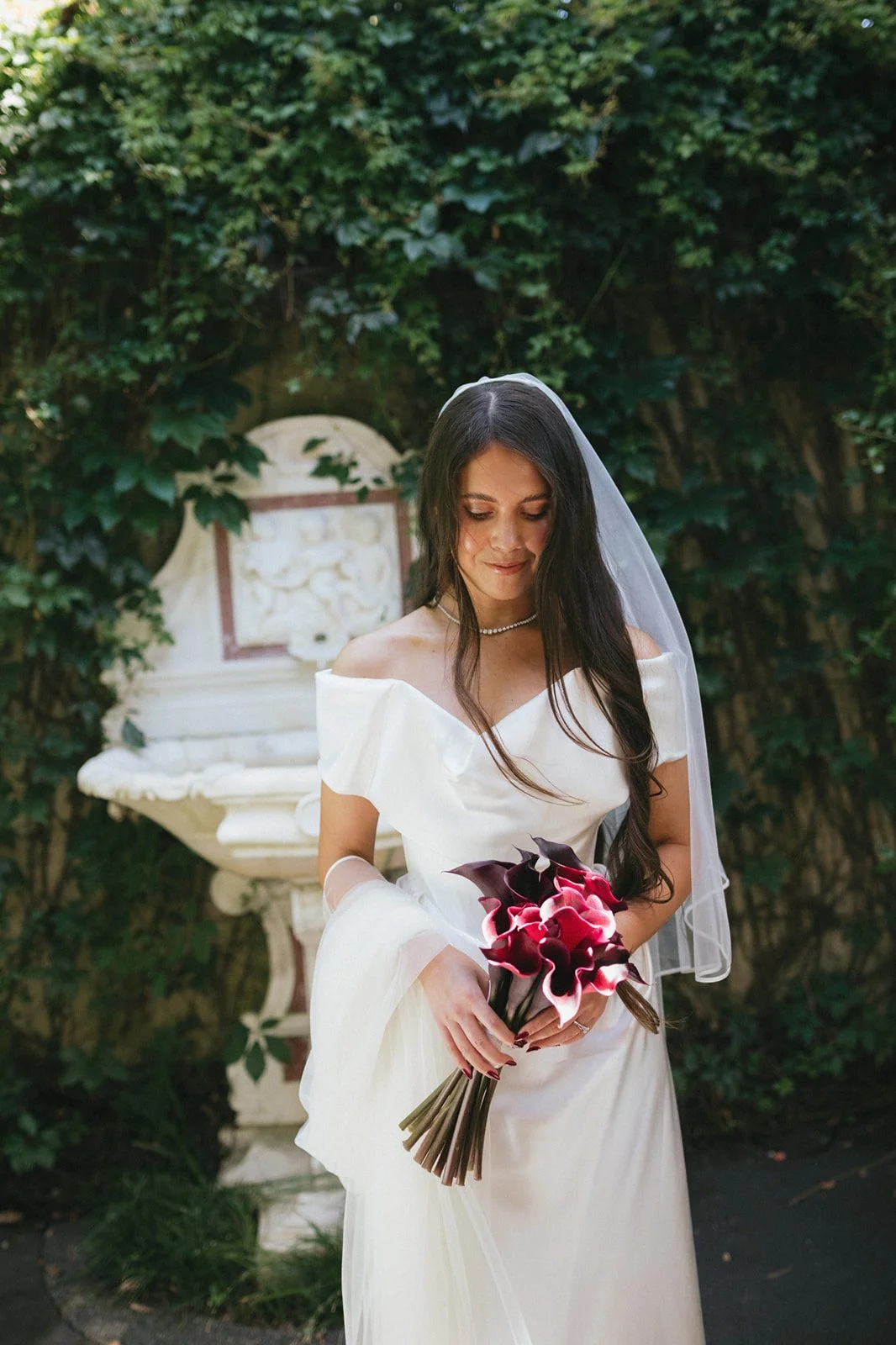 A bride in a white wedding dress holding a bouquet of dark pink and Burgundy calla lilies, standing outdoors with greenery and a decorative stone fountain in the background.