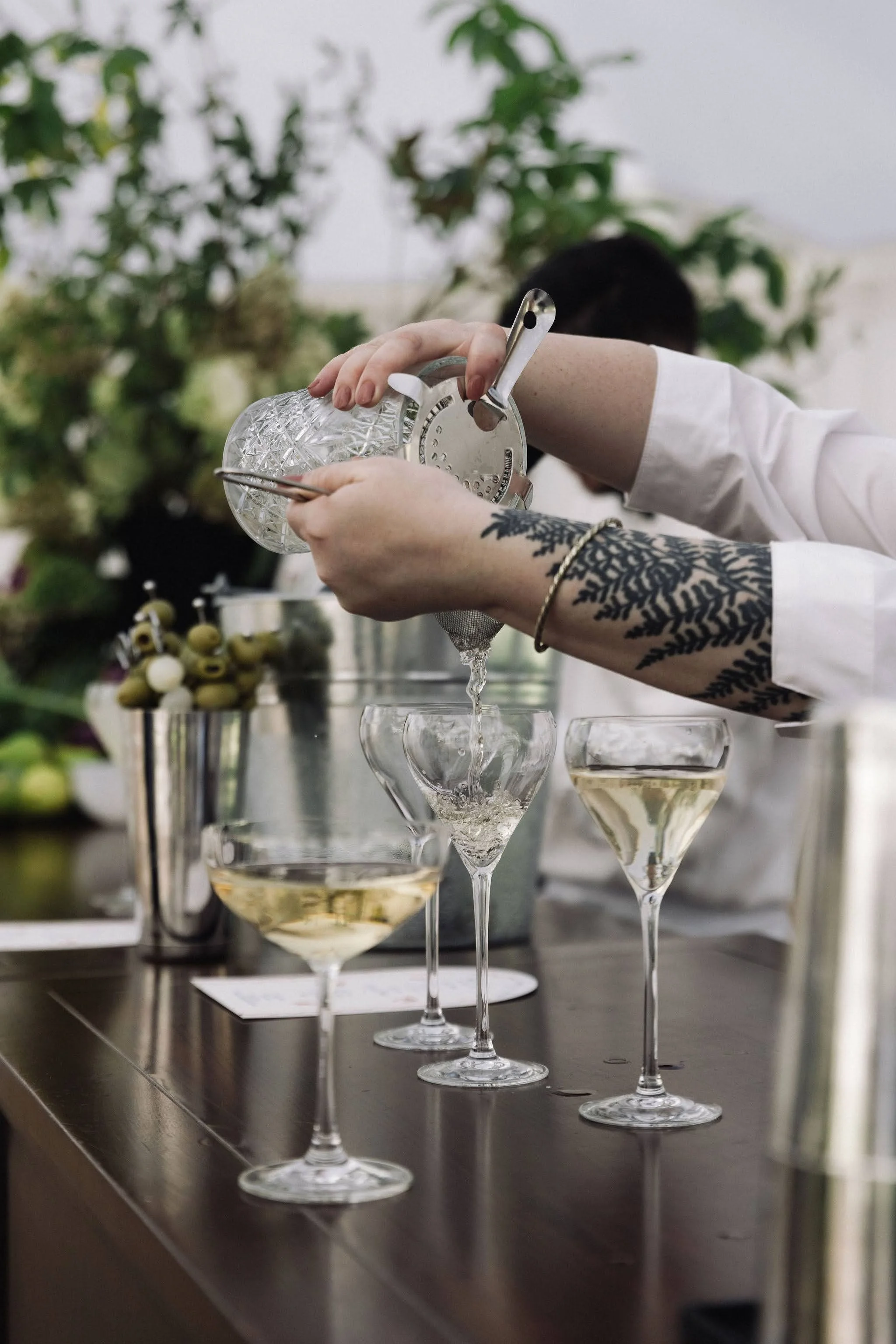 A person with a tattoo on their arm pouring a drink into a martini glass at a bar or event, with other filled glasses and a floral arrangement in the background.