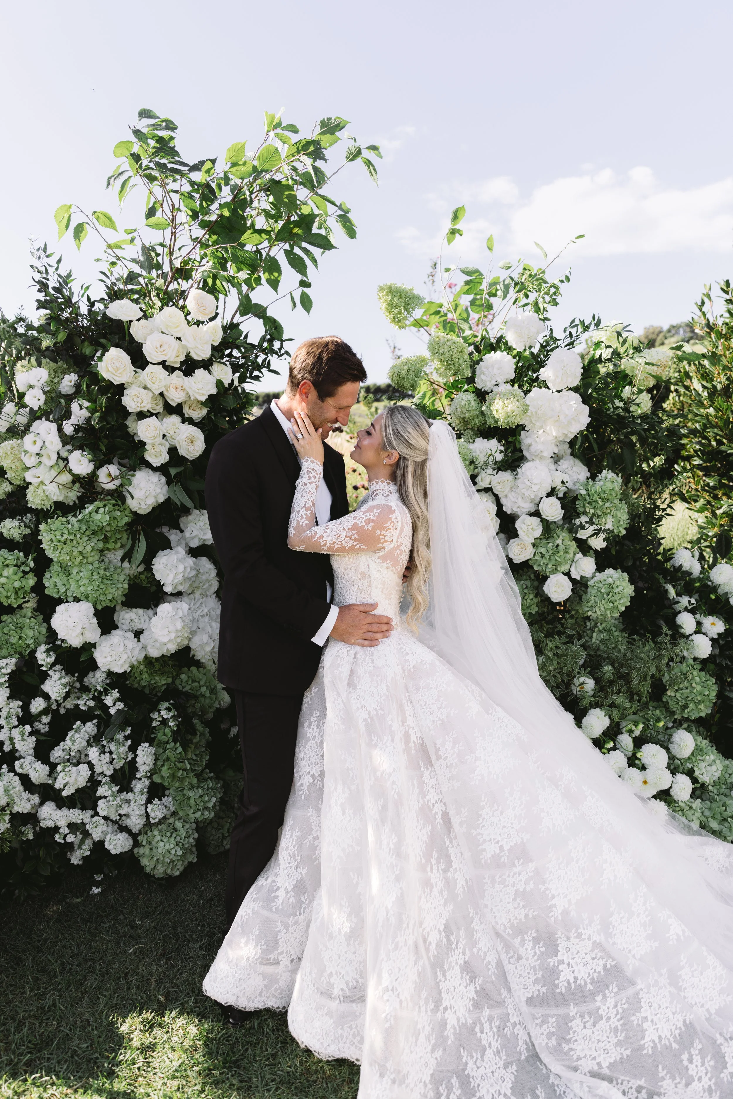 A bride and groom sharing a kiss outdoors surrounded by white roses and greenery during a wedding.