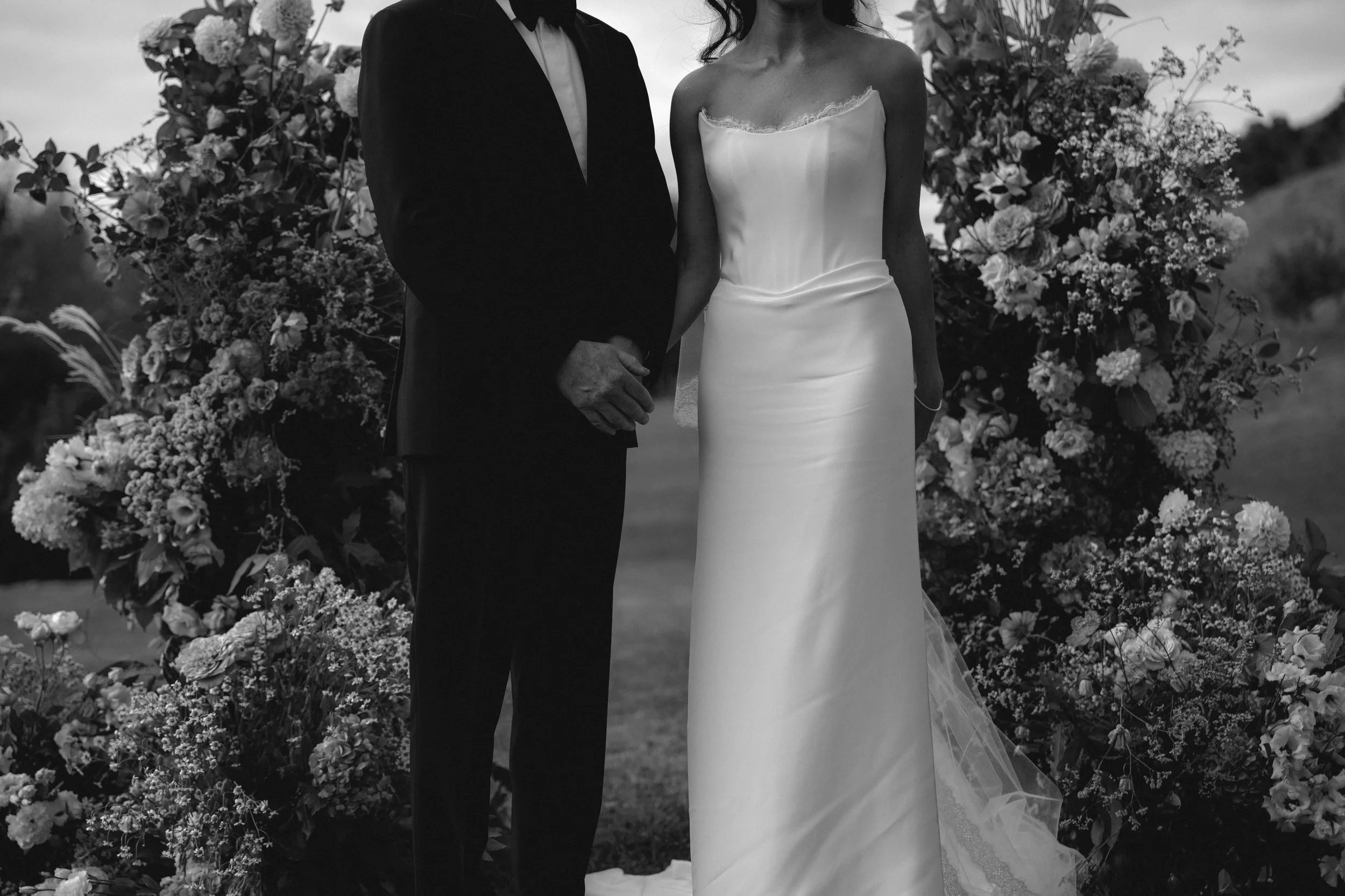 A black and white photo of a bride and groom holding hands during a wedding ceremony, surrounded by floral arrangements.