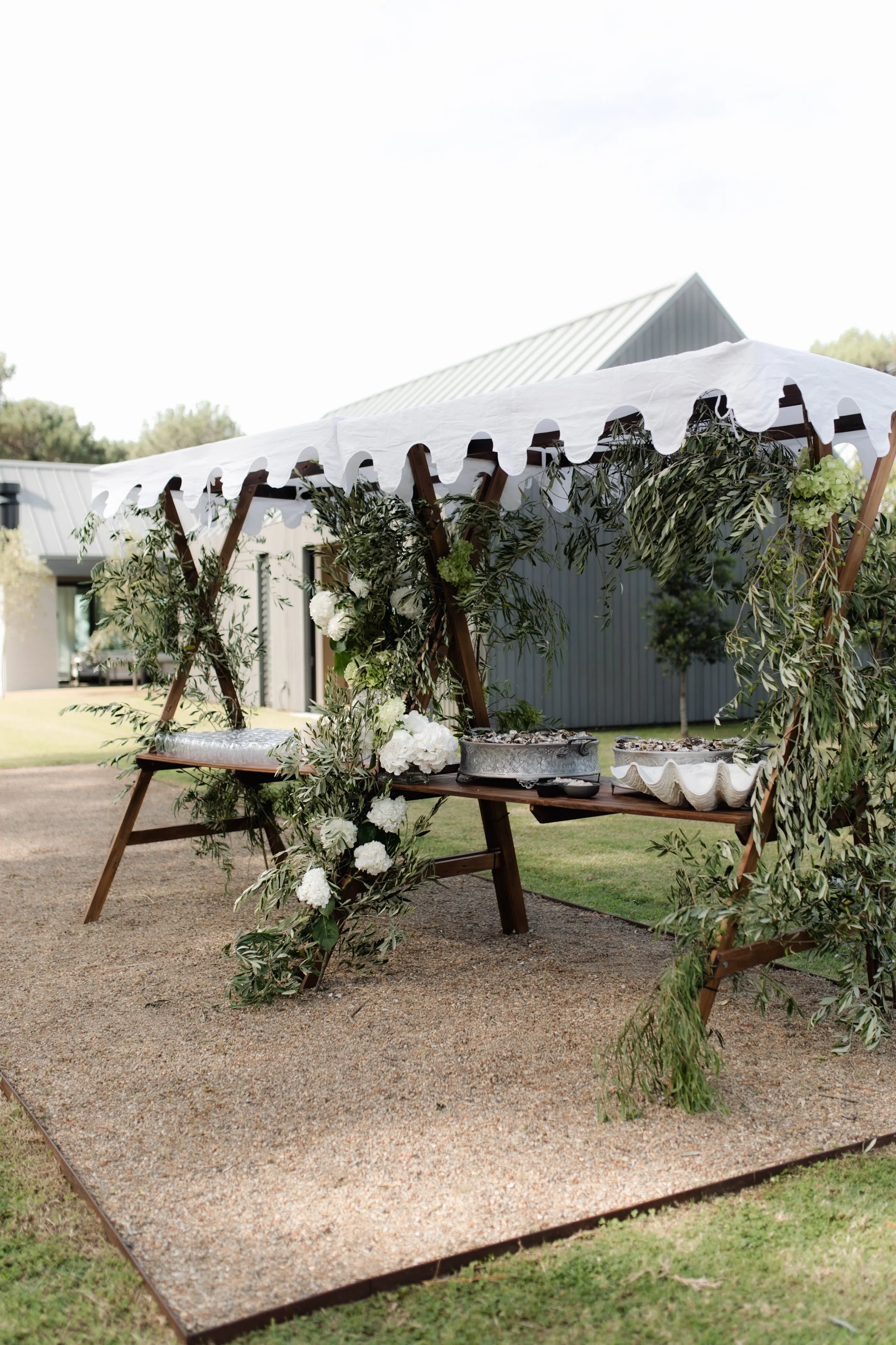Wedding outdoor setup with a decorated wooden arch covered in greenery and white flowers, with a white scalloped canopy, on a gravel platform in a grassy area, likely for a ceremony.
