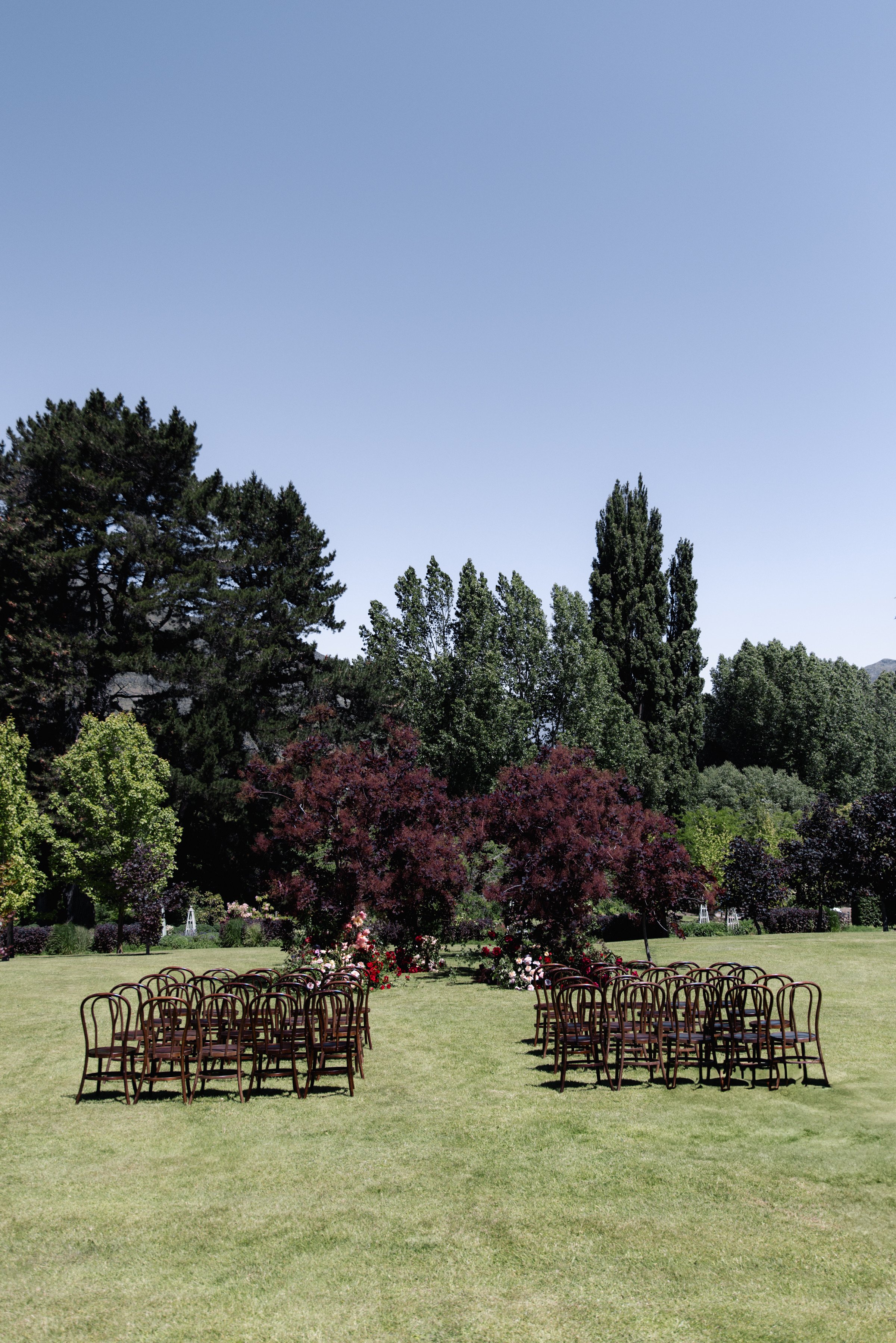 Outdoor wedding ceremony setup with rows of chairs on a grassy field, surrounded by colorful trees and flower arrangements, with a large blue sky overhead.
