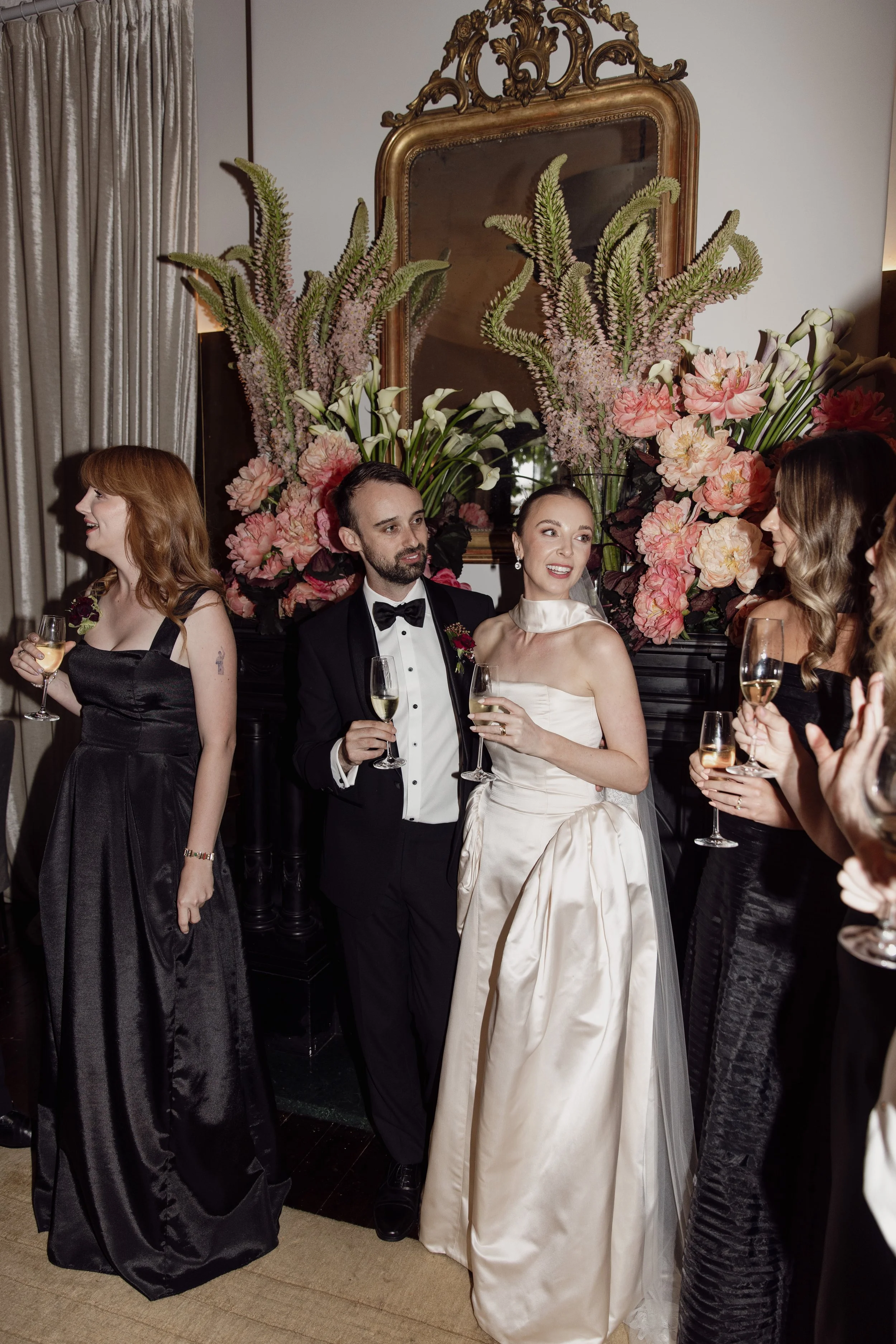 People dressed in formal attire at a celebration, holding glasses of champagne, amidst large floral arrangements with pink and green flowers, in a room with a vintage mirror and cream-colored curtains.