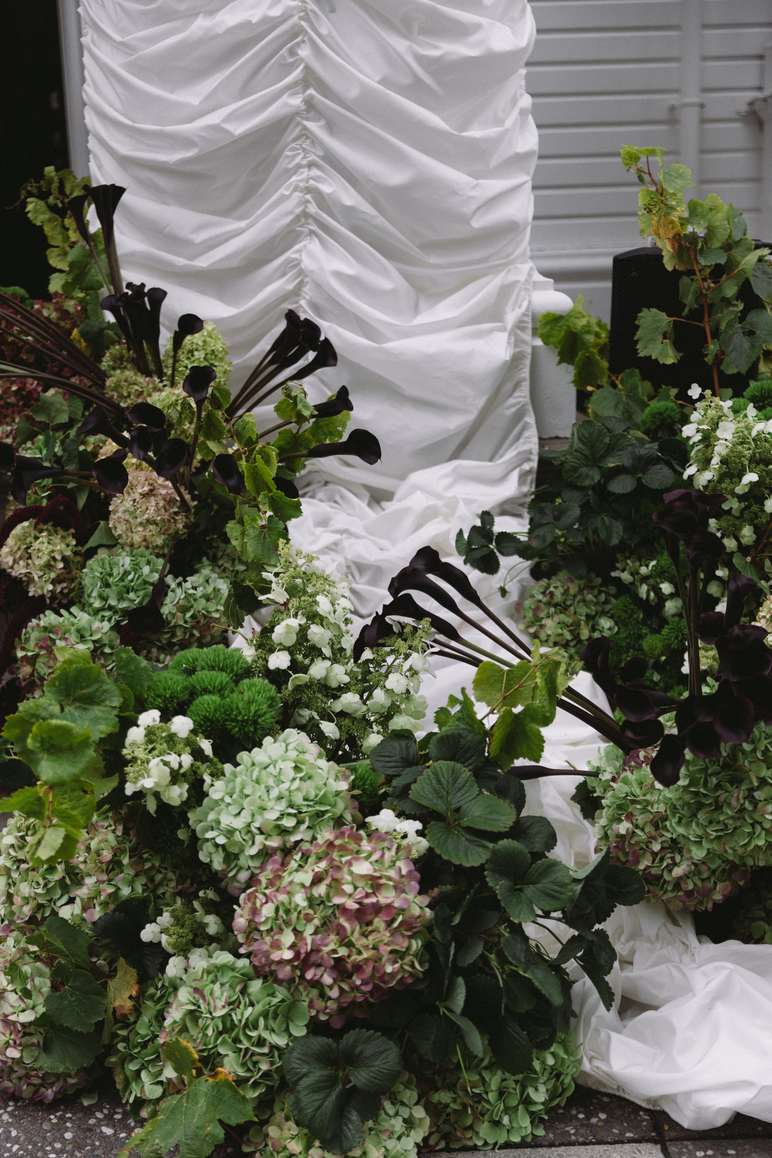 A large bouquet of colorful flowers with dark purple, green, and white blooms, placed next to a white fabric backdrop or drapery.