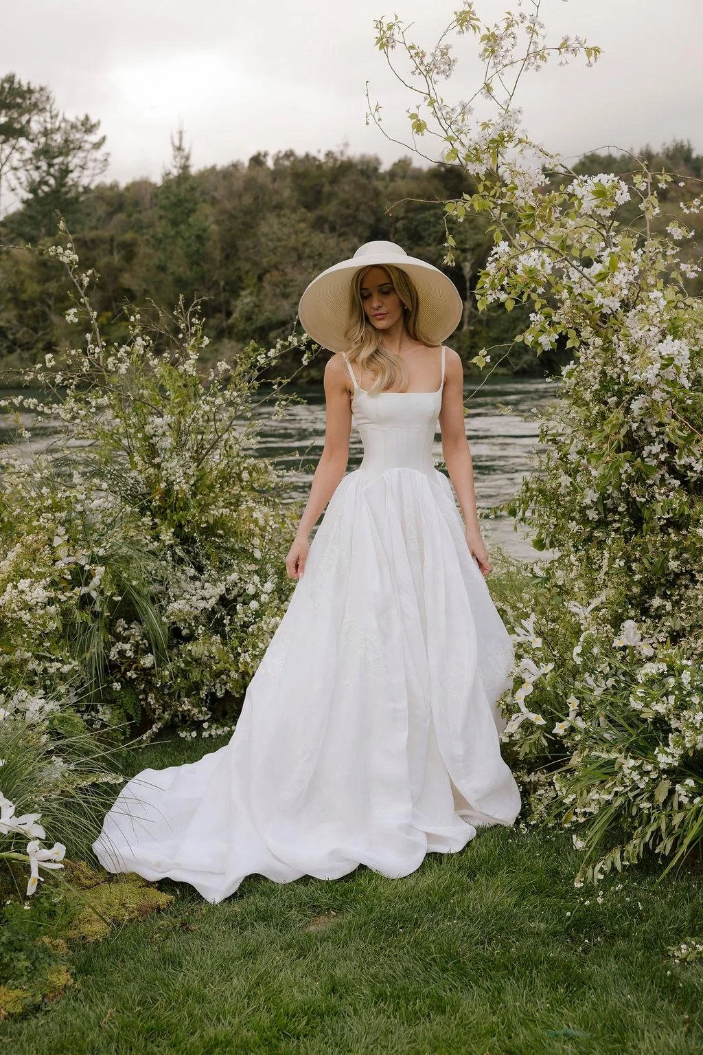 A bride in a white wedding dress with a wide-brimmed hat standing among blooming white flowers near a body of water and trees in the background at Huka Lodge, Taupo