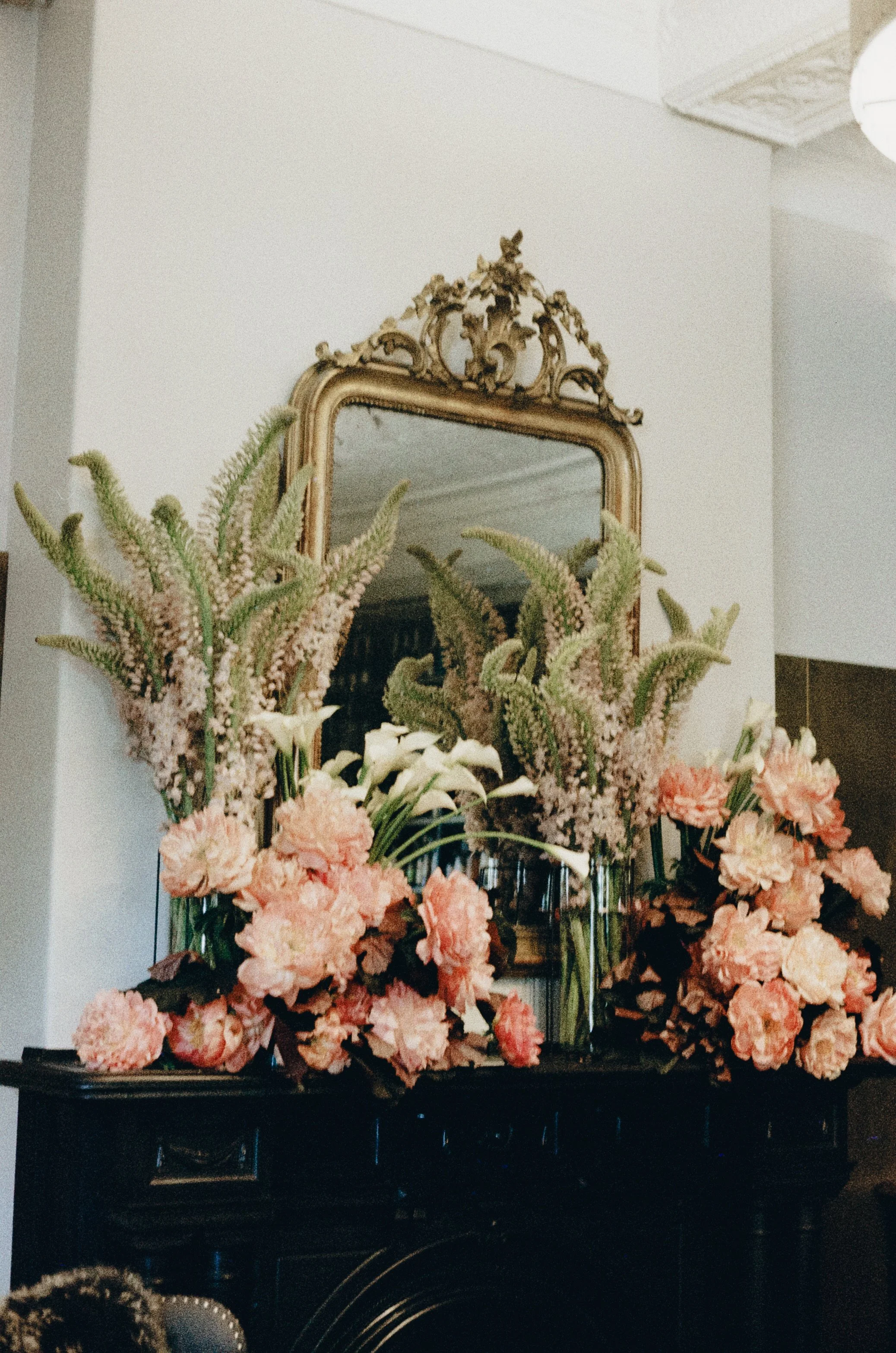A decorative mirror with an ornate gold frame is placed on a wall. In front of the mirror, there are numerous pink and white flowers arranged in vases, creating a floral display in an indoor setting.