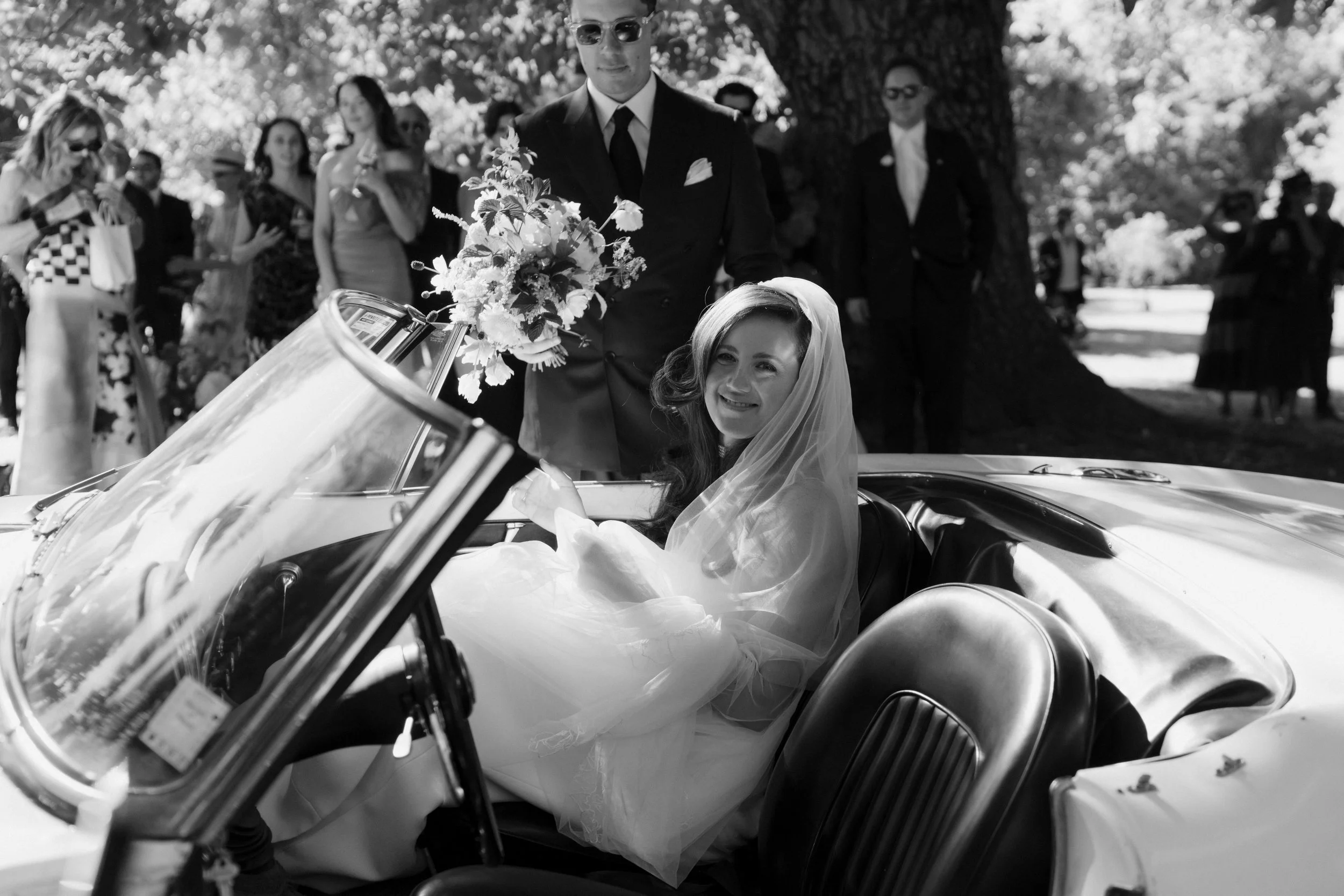 A bride in a wedding dress and veil sitting in a vintage convertible car, smiling. A man in a suit and sunglasses is holding a bouquet of flowers and standing beside the car. Several guests are gathered outdoors under trees, some taking photos, dressed in formal and semi-formal attire.