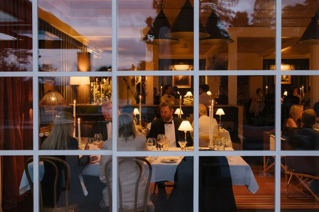 People dining at a restaurant seen through a window with grid panes, with candlelit tables and warm lighting inside.